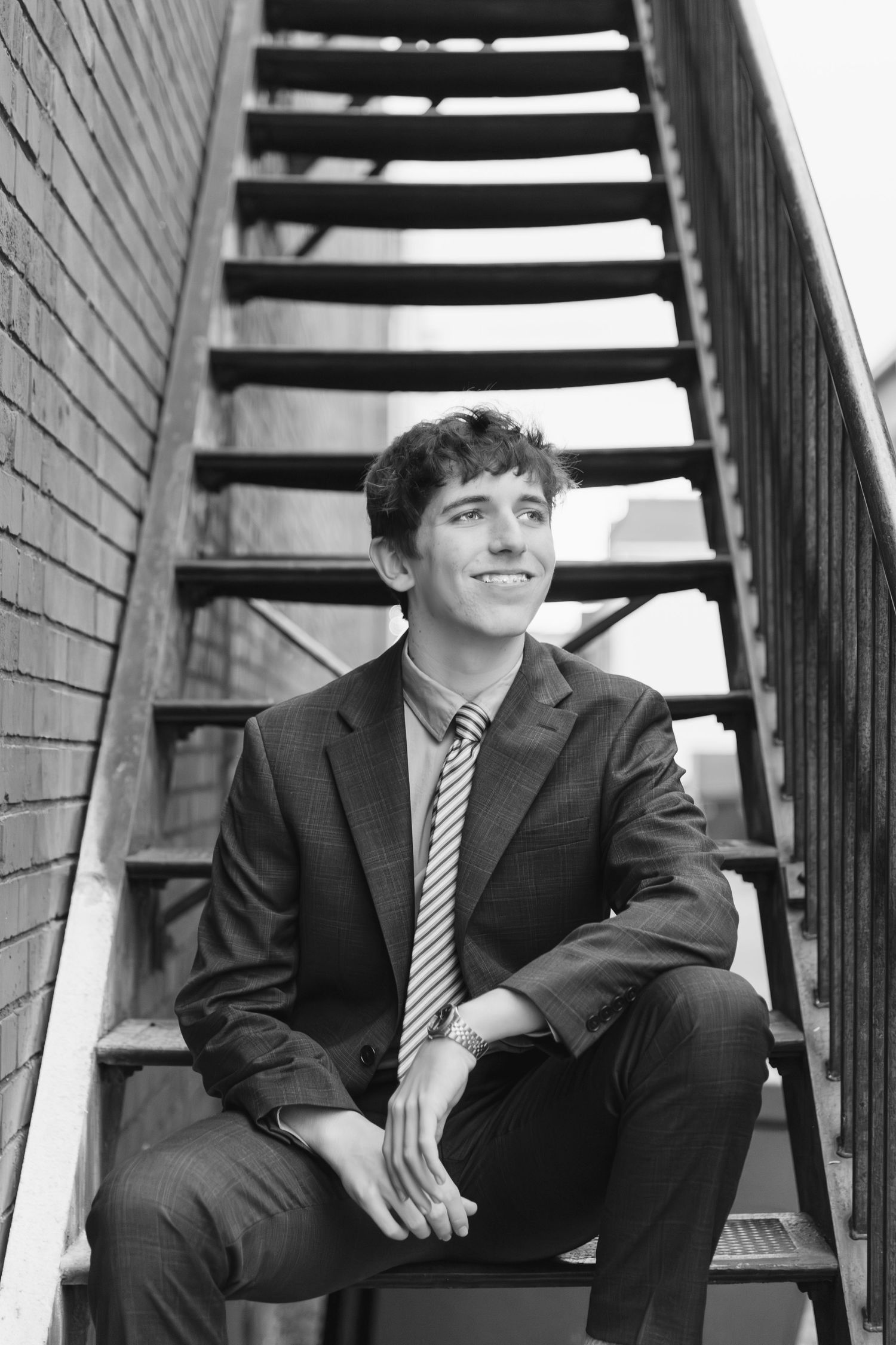 High school senior boy in formal attire poses on metal staircase in black and white photo in Downtown Bartlesville, Oklahoma.