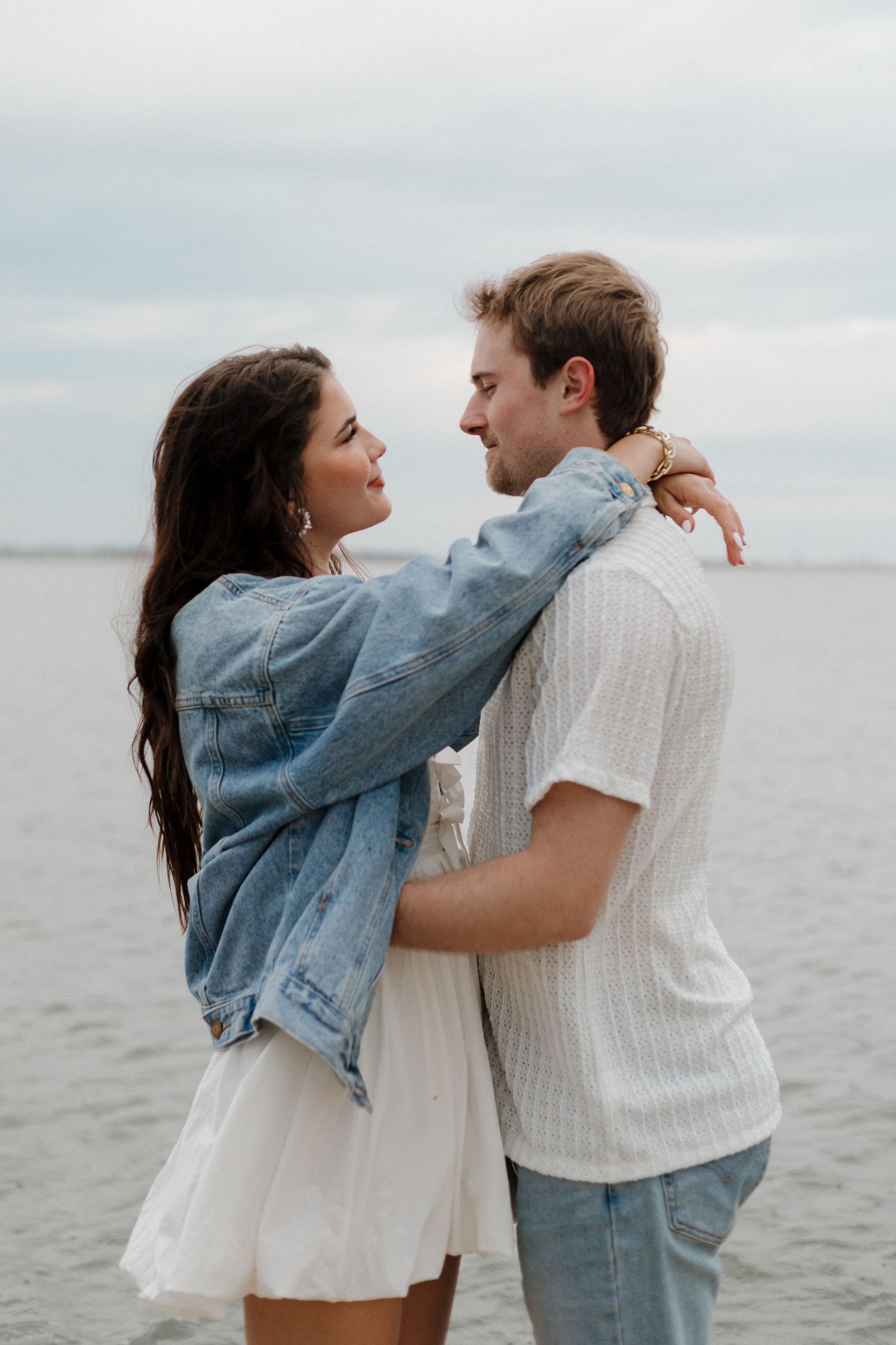 A romantic couple embraces by the water wearing casual denim and white clothing.