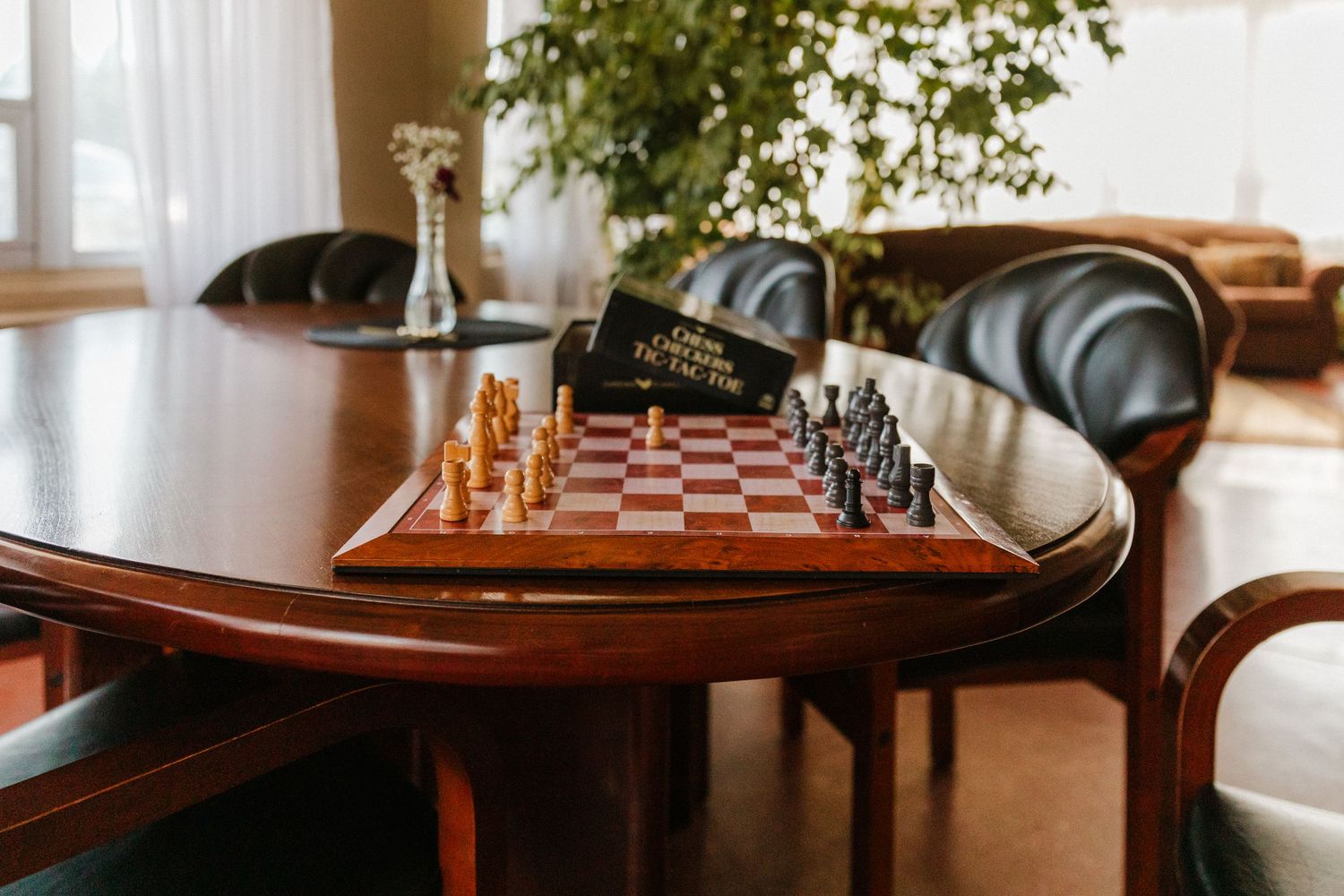 A wooden chess board and pieces sit on an elegant table near a window.