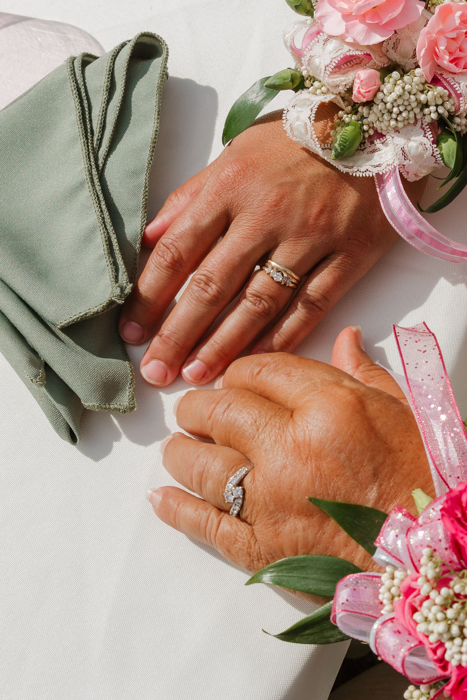 Close-up of hands wearing wedding rings surrounded by pink flowers and greenery.