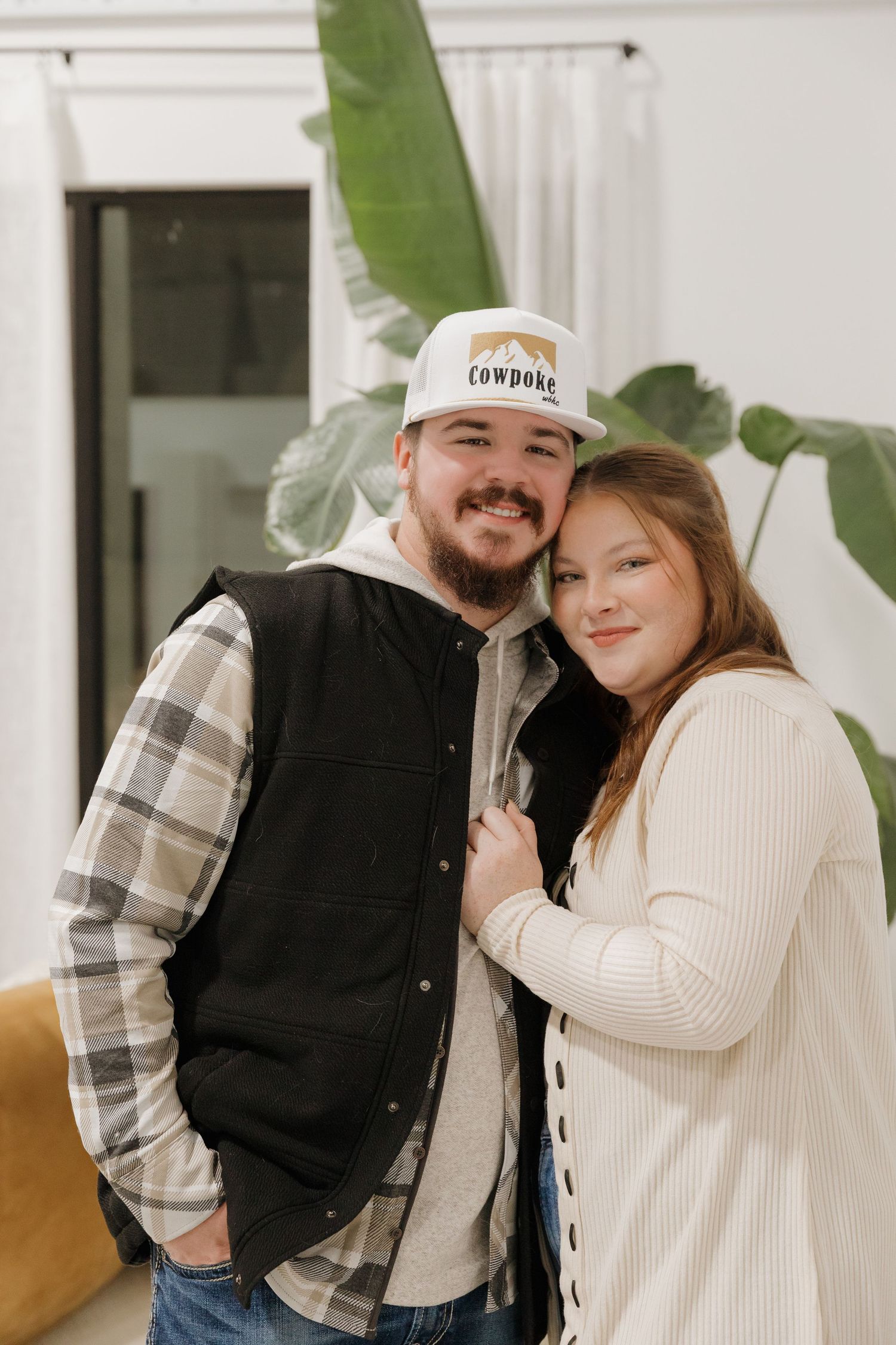 Couple in casual winter wear posing against white wall with tropical plant.