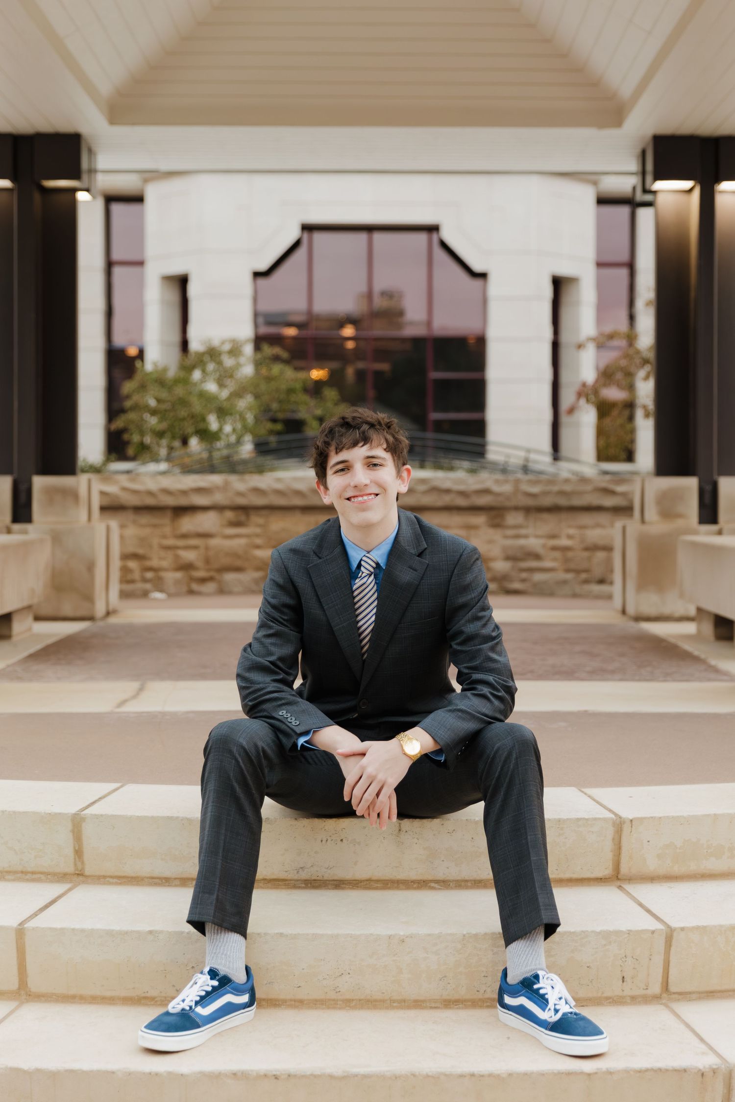 Person in formal suit and blue sneakers sits on concrete steps outside building.