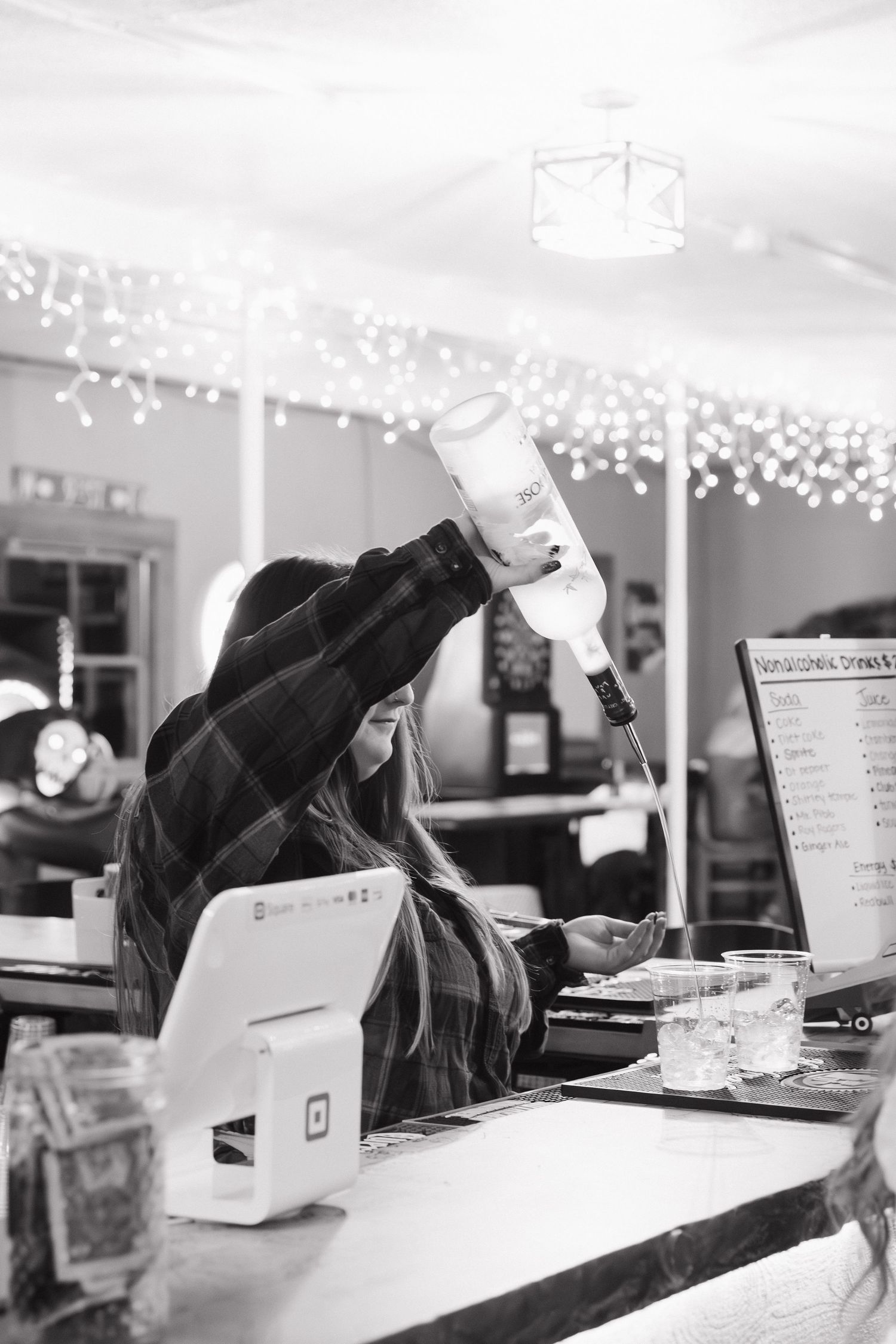 Black and white photo of bartender at illuminated counter with string lights above at a venue in Volga, South Dakota.