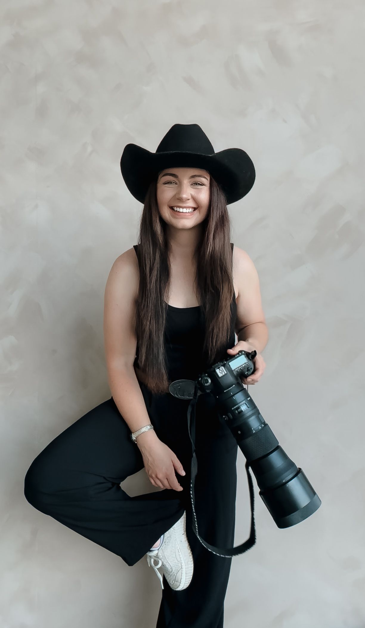 Professional photographer in black dress and cowboy hat posing with camera equipment.