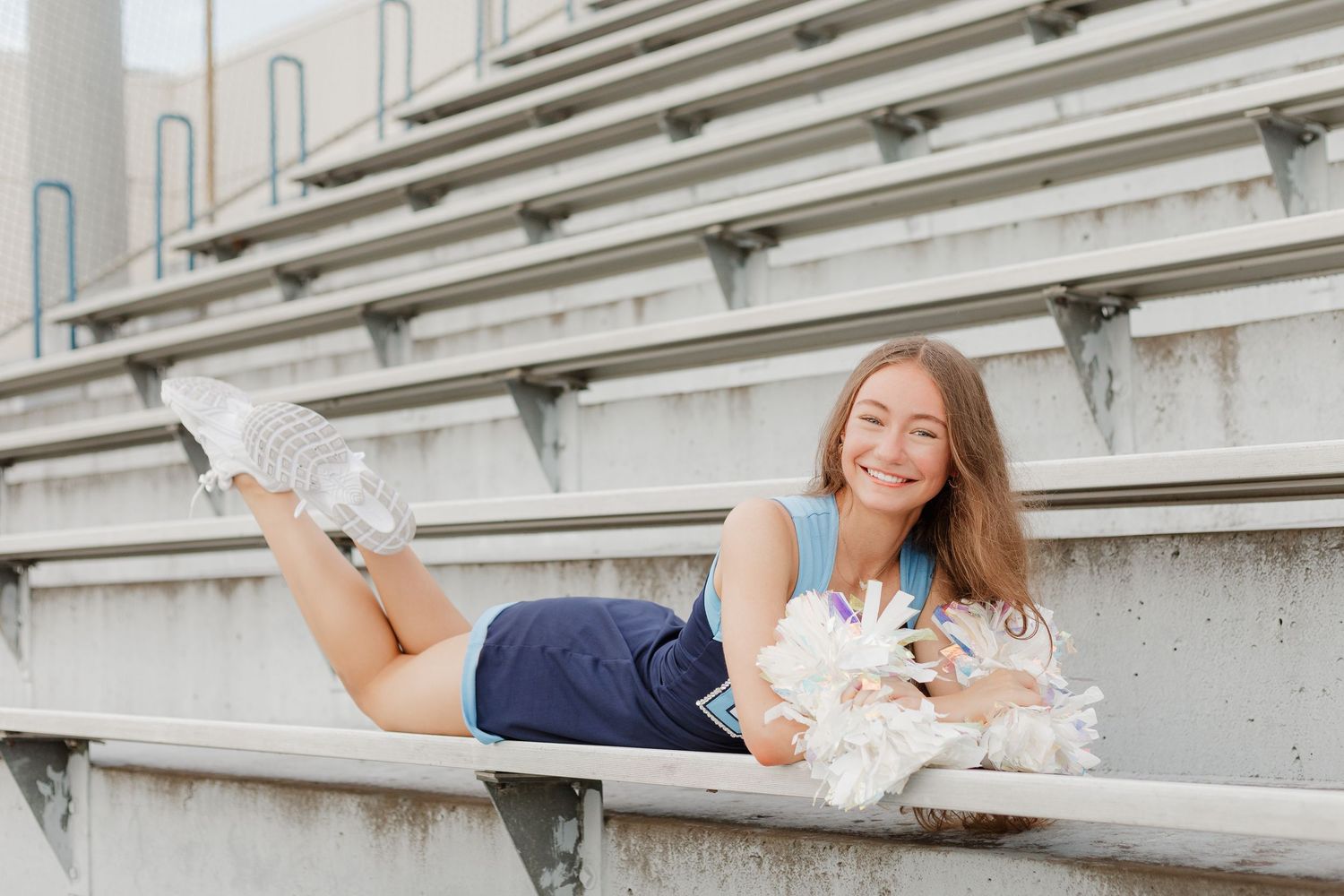 Cheerleader relaxing on stadium bleachers with white pom poms in casual pose.
