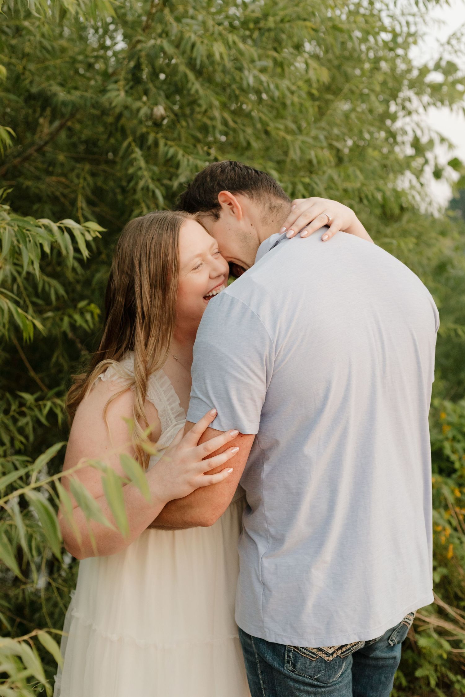 A romantic couple embraces against a backdrop of lush green foliage in a natural outdoor setting.
