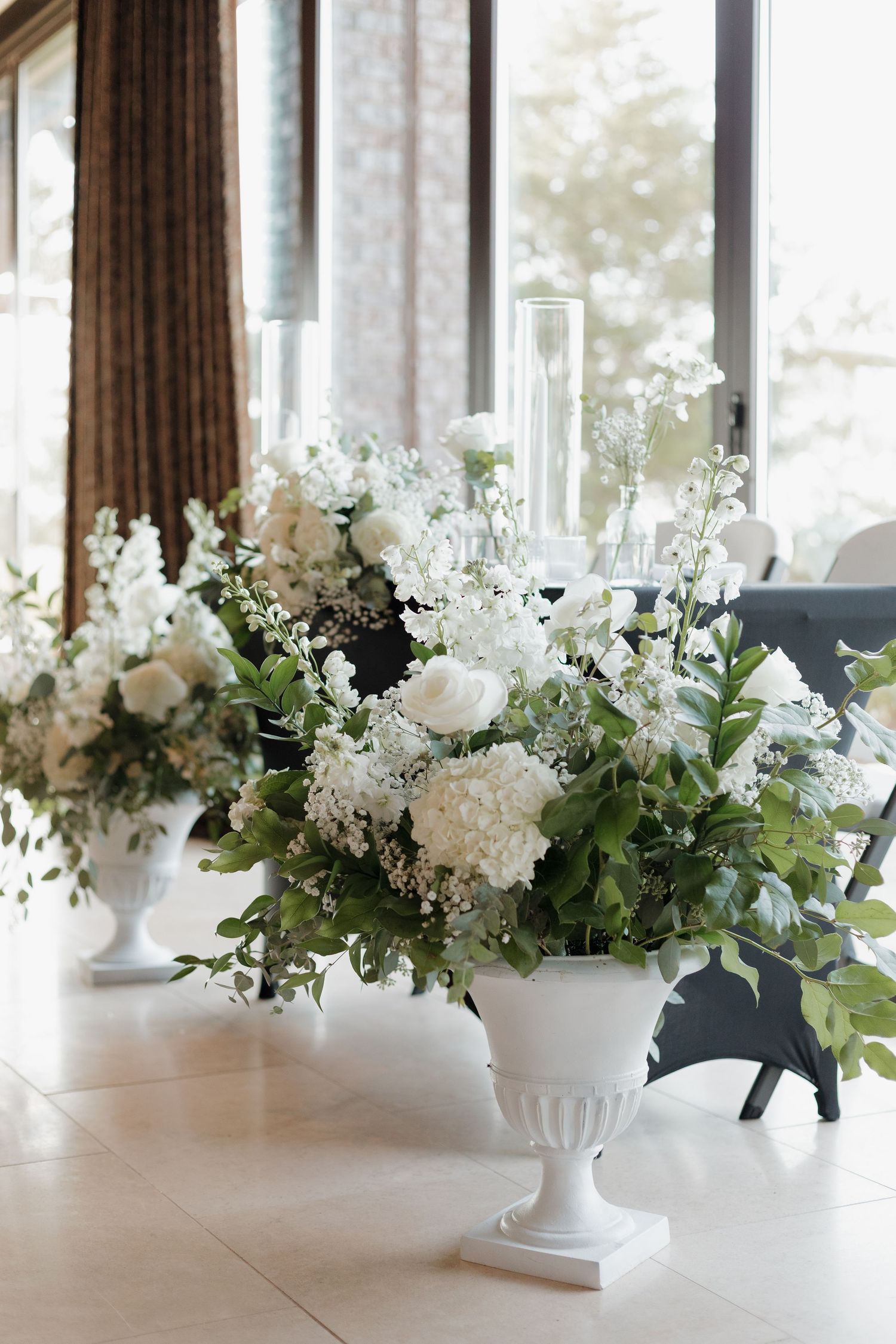 Elegant white floral centerpieces with roses and greenery arranged in white pedestal vases on a long table.