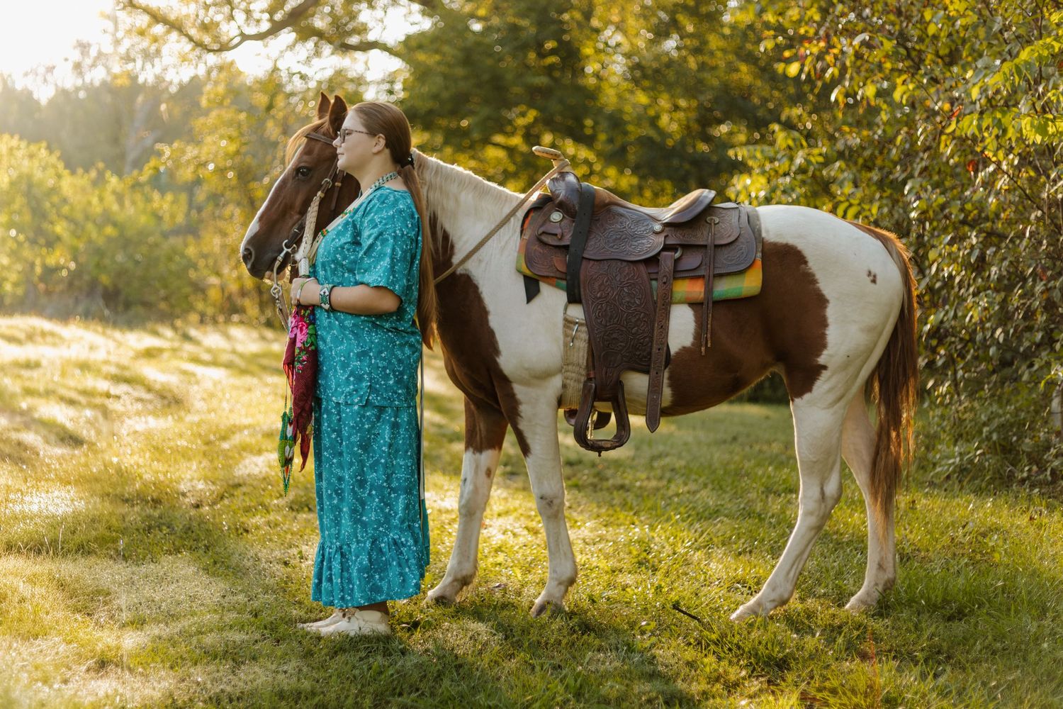 Paint horse and rider in turquoise dress standing in golden sunlit field.