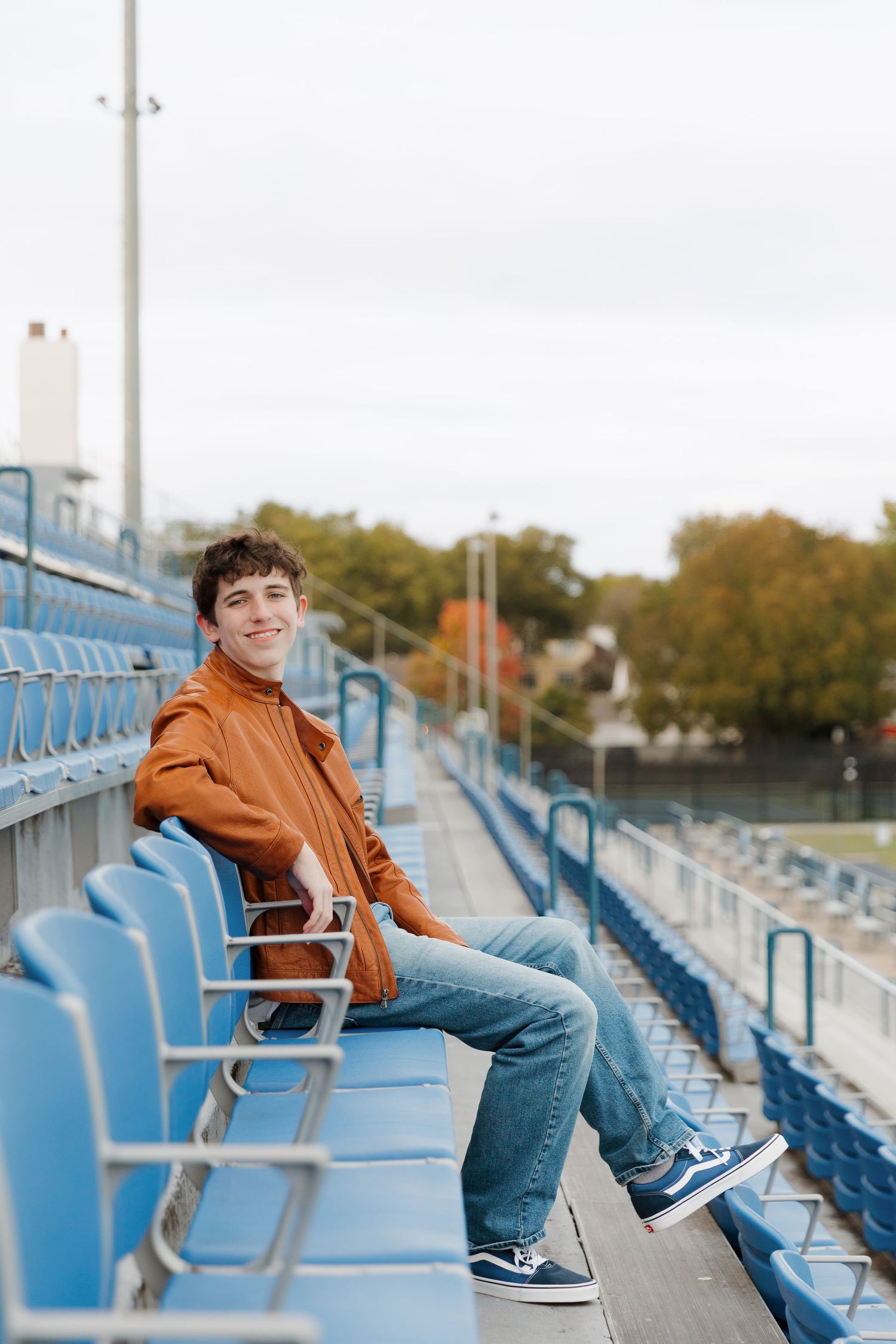 Person relaxes in blue stadium seats wearing brown jacket and jeans.