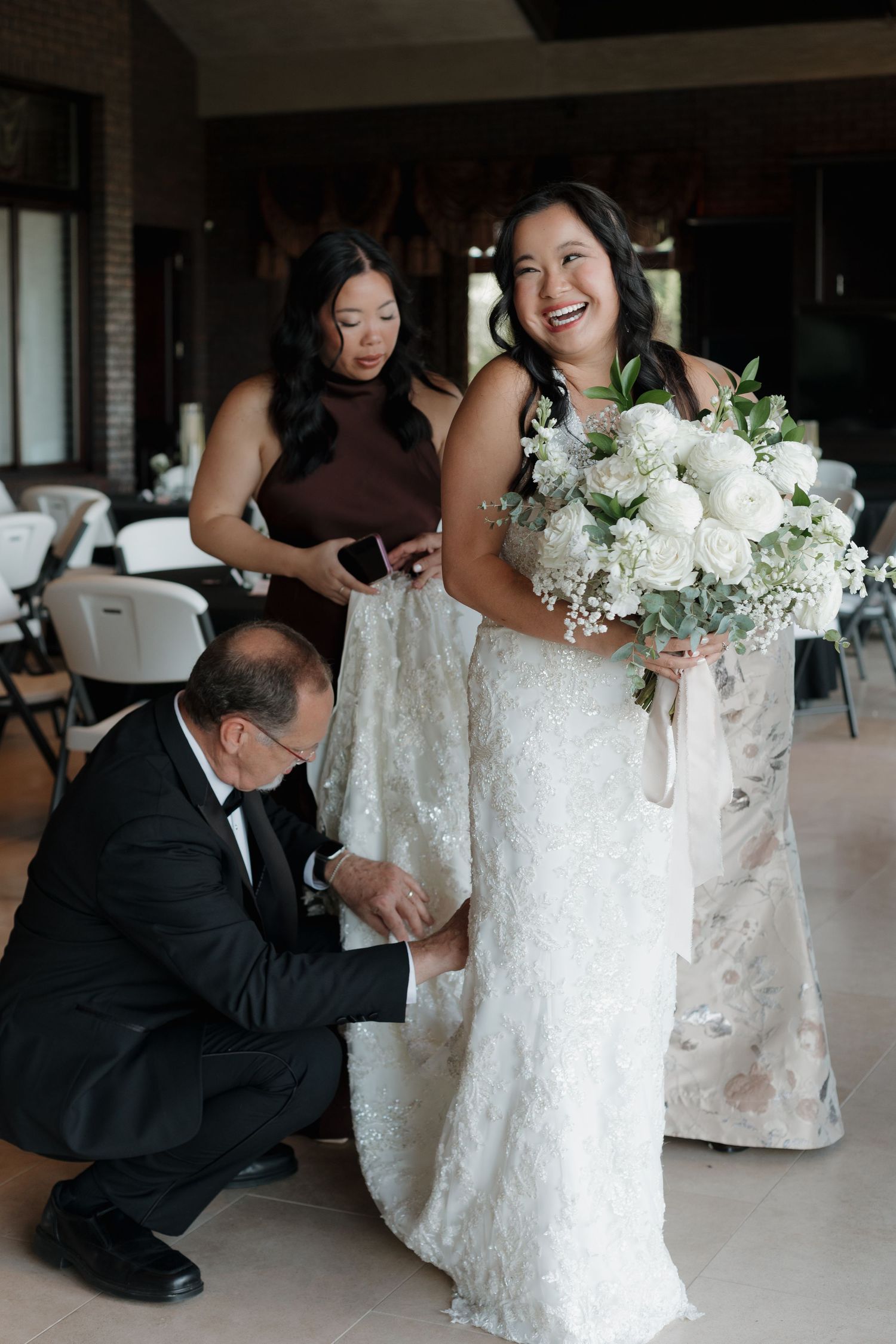 Wedding preparations showing people adjusting a white lace dress in an indoor venue.