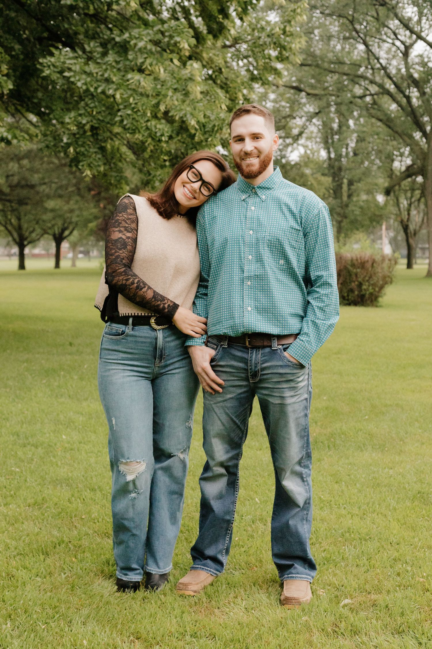 A couple in casual attire standing together on a grassy lawn with trees in the background.