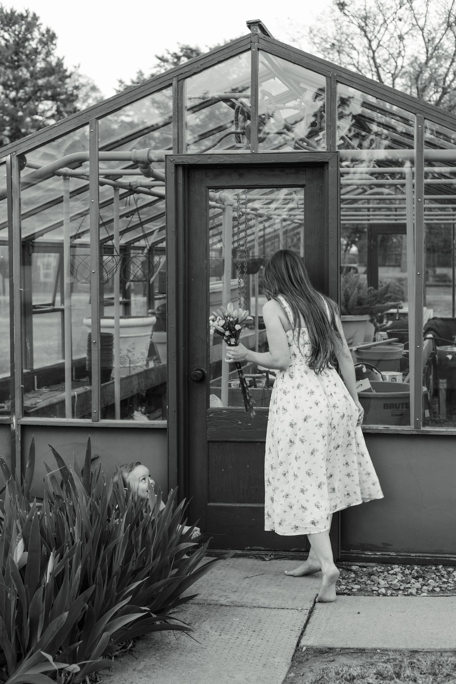 Person in floral dress explores greenhouse exterior in black and white photo.