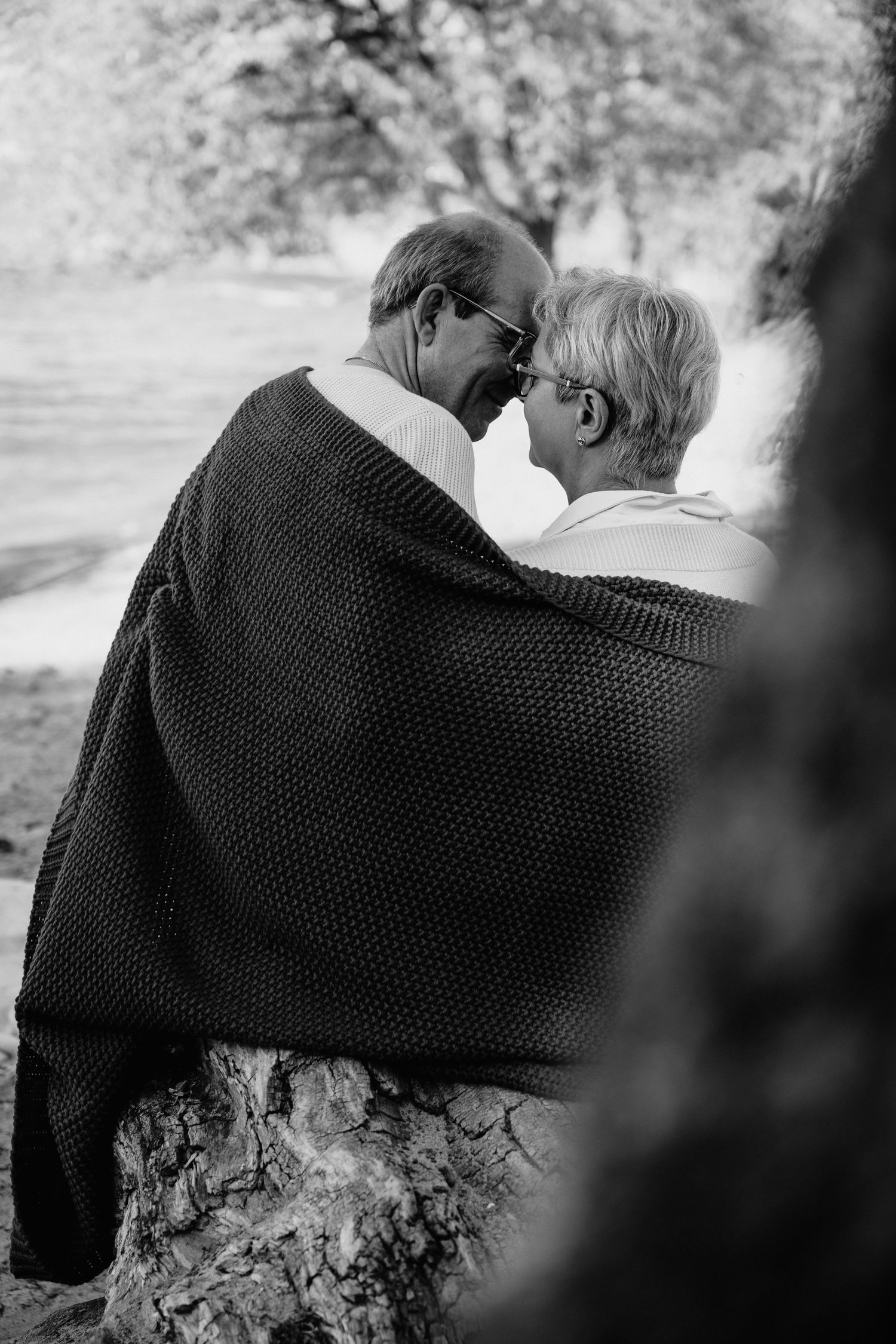 Black and white photo of an elderly couple sharing an intimate moment, wrapped in a blanket outdoors.