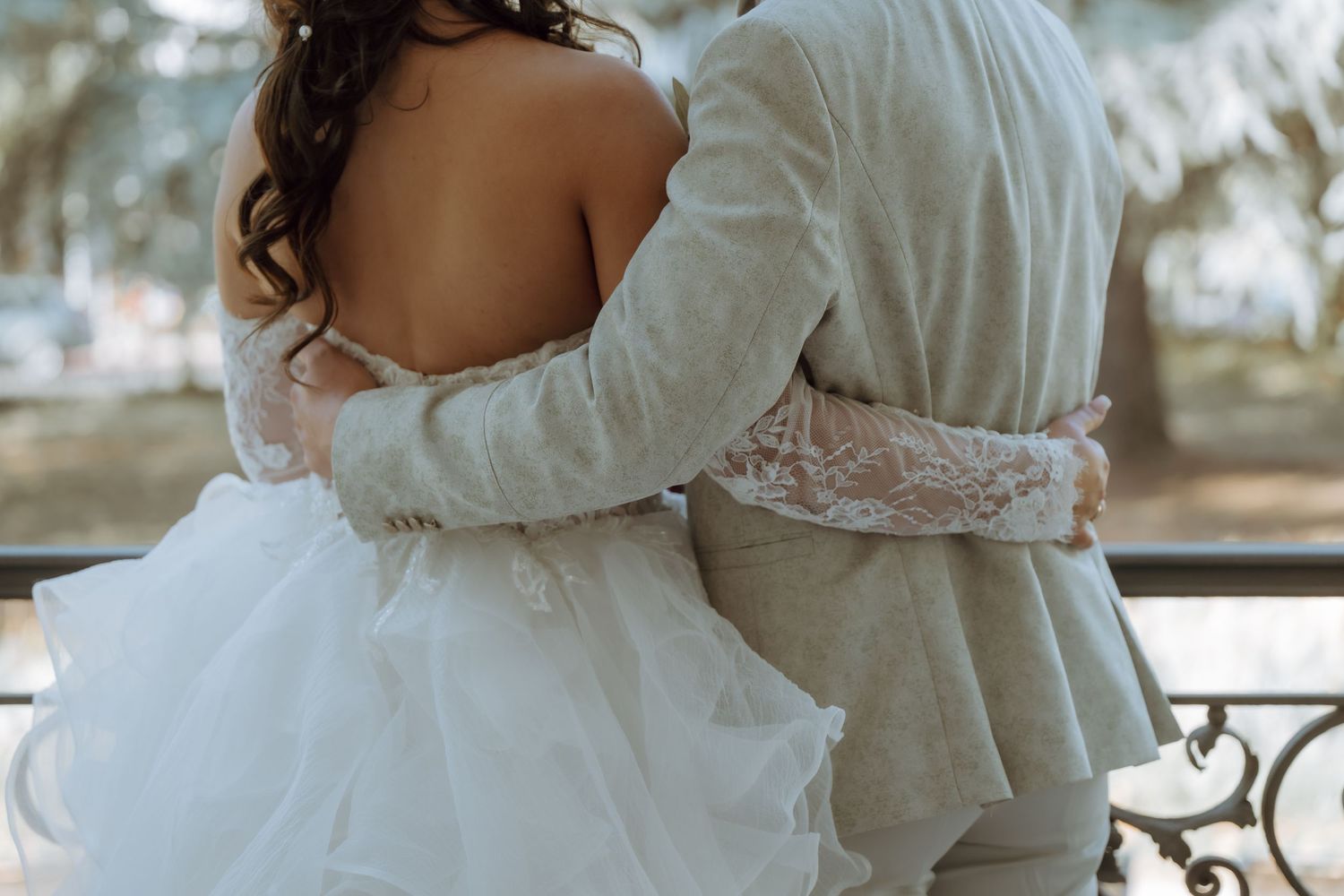A bride and groom embrace from behind, showcasing her lace wedding dress and his gray suit outdoors.