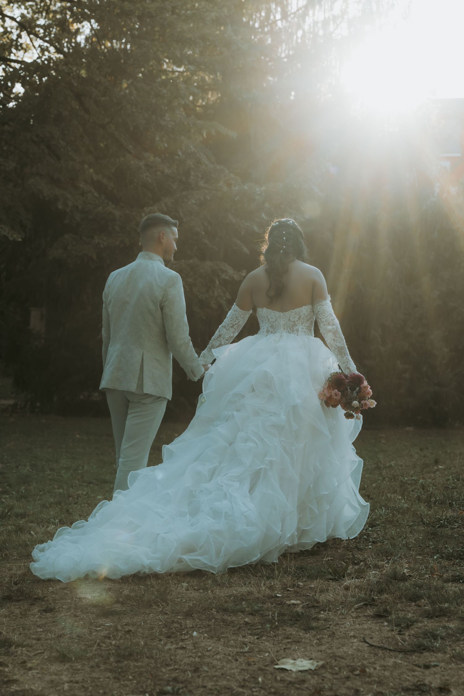 Bride and groom walking hand in hand toward sunlit trees, bride in ruffled white gown holding bouquet.
