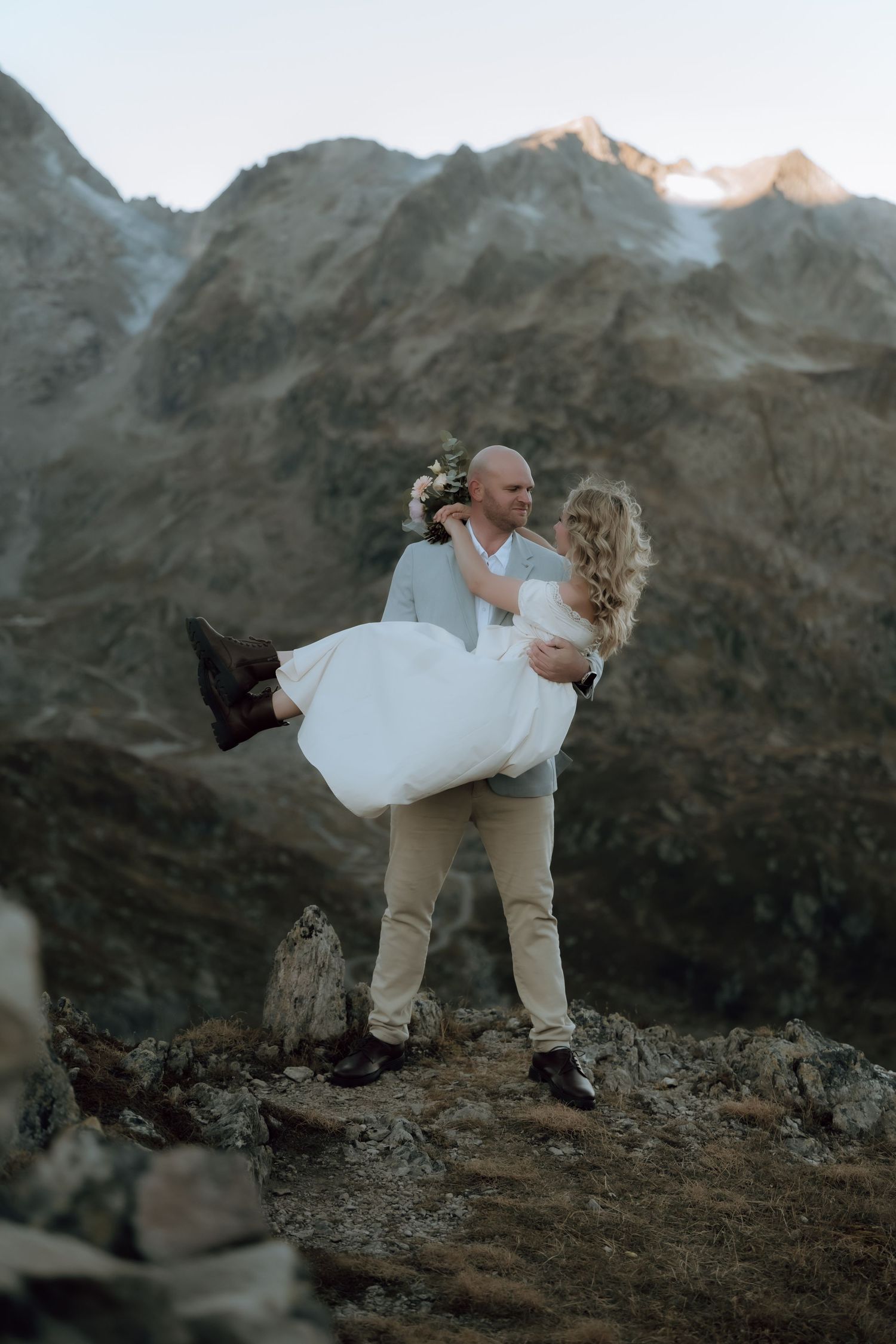 Groom carries bride in white dress on rocky mountain summit during outdoor wedding elopement at dusk.