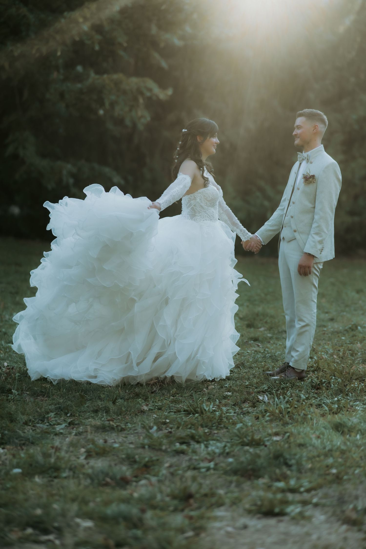 Bride in flowing white gown holds hands with groom in beige suit, dancing in sunlit garden.