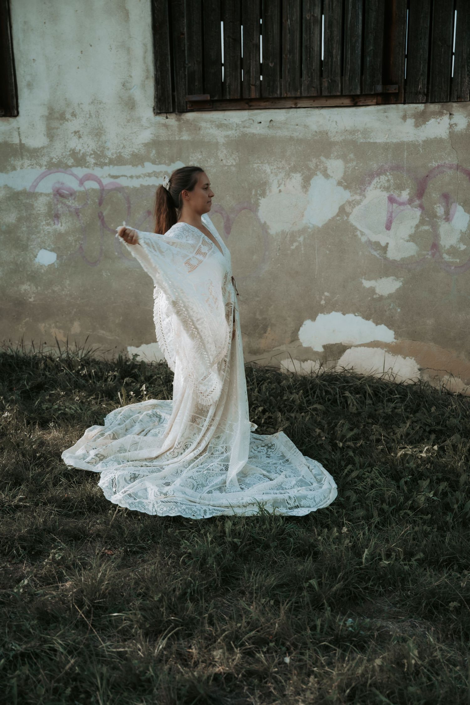 Woman in flowing white lace bridal gown posing outdoors near a weathered wall with graffiti.
