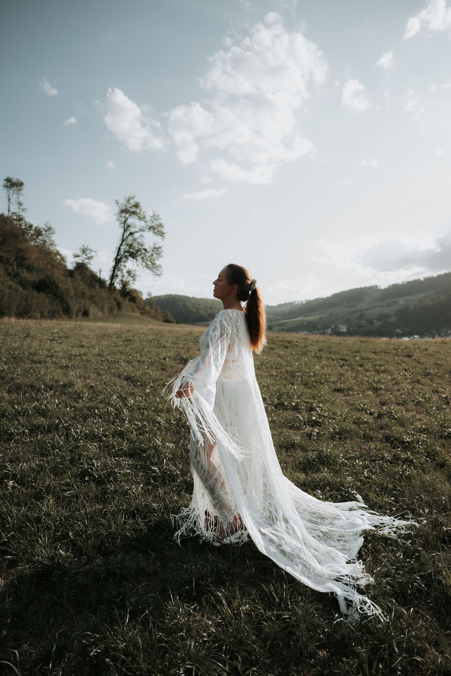 Bride in flowing white lace gown standing in green meadow with rolling hills and cloudy sky backdrop.