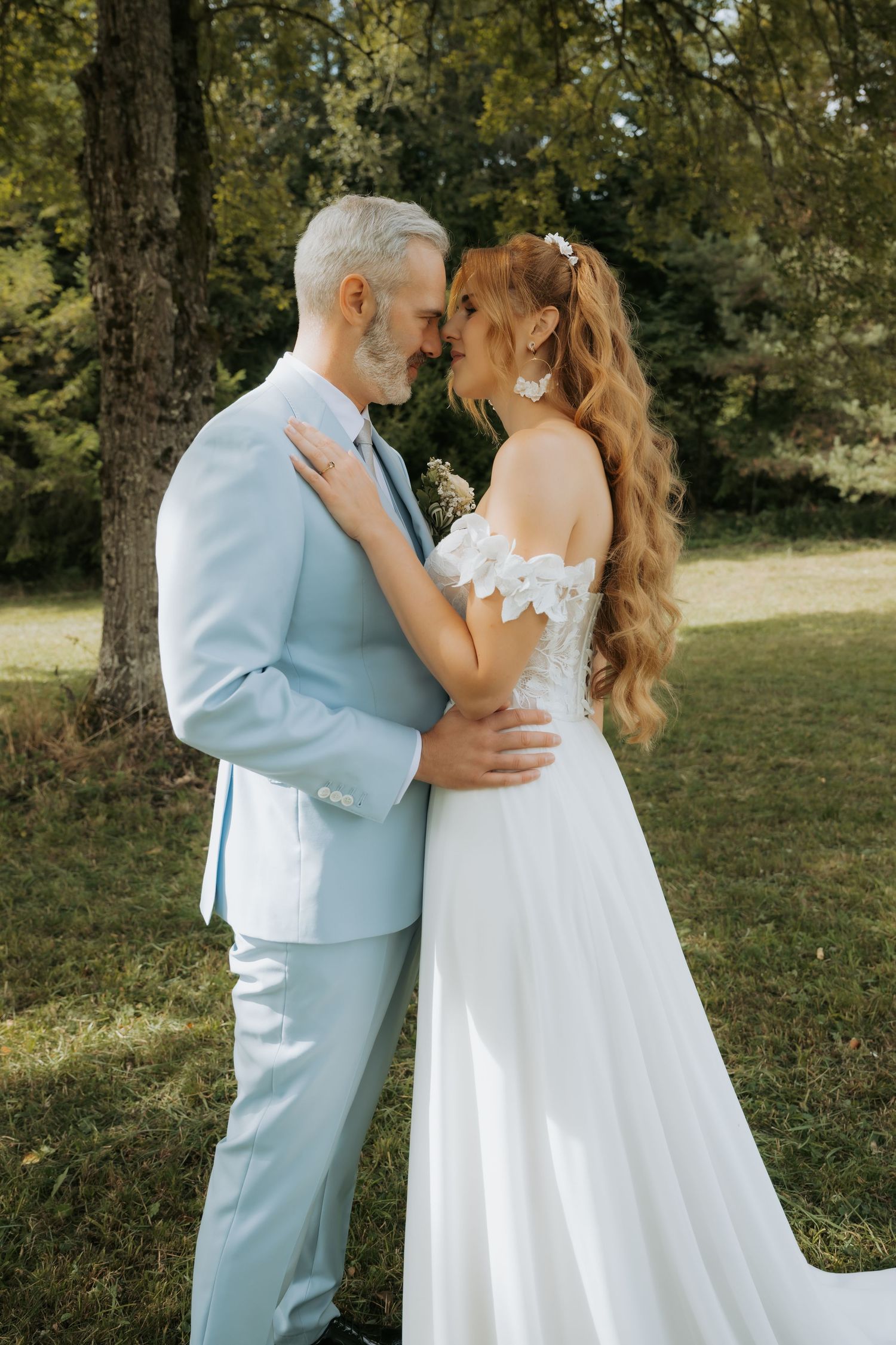 A bride and groom share a romantic moment outdoors, with the groom in a light blue suit and bride in a white lace gown.