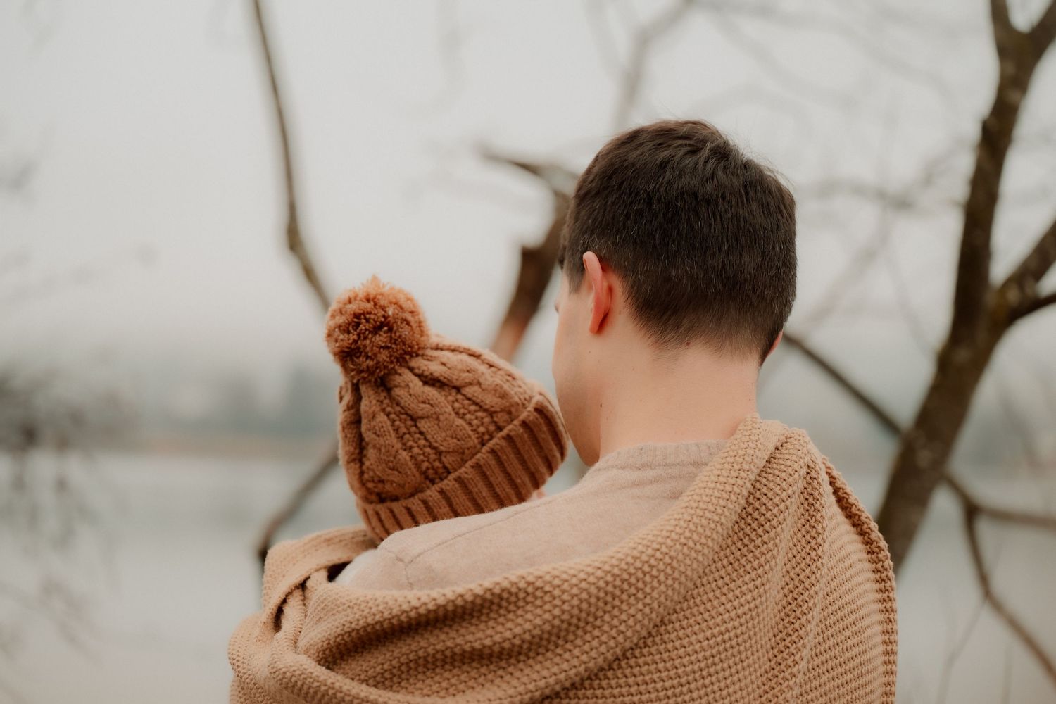 A man holds a child wearing a brown knit pom-pom hat outdoors near bare trees in winter.