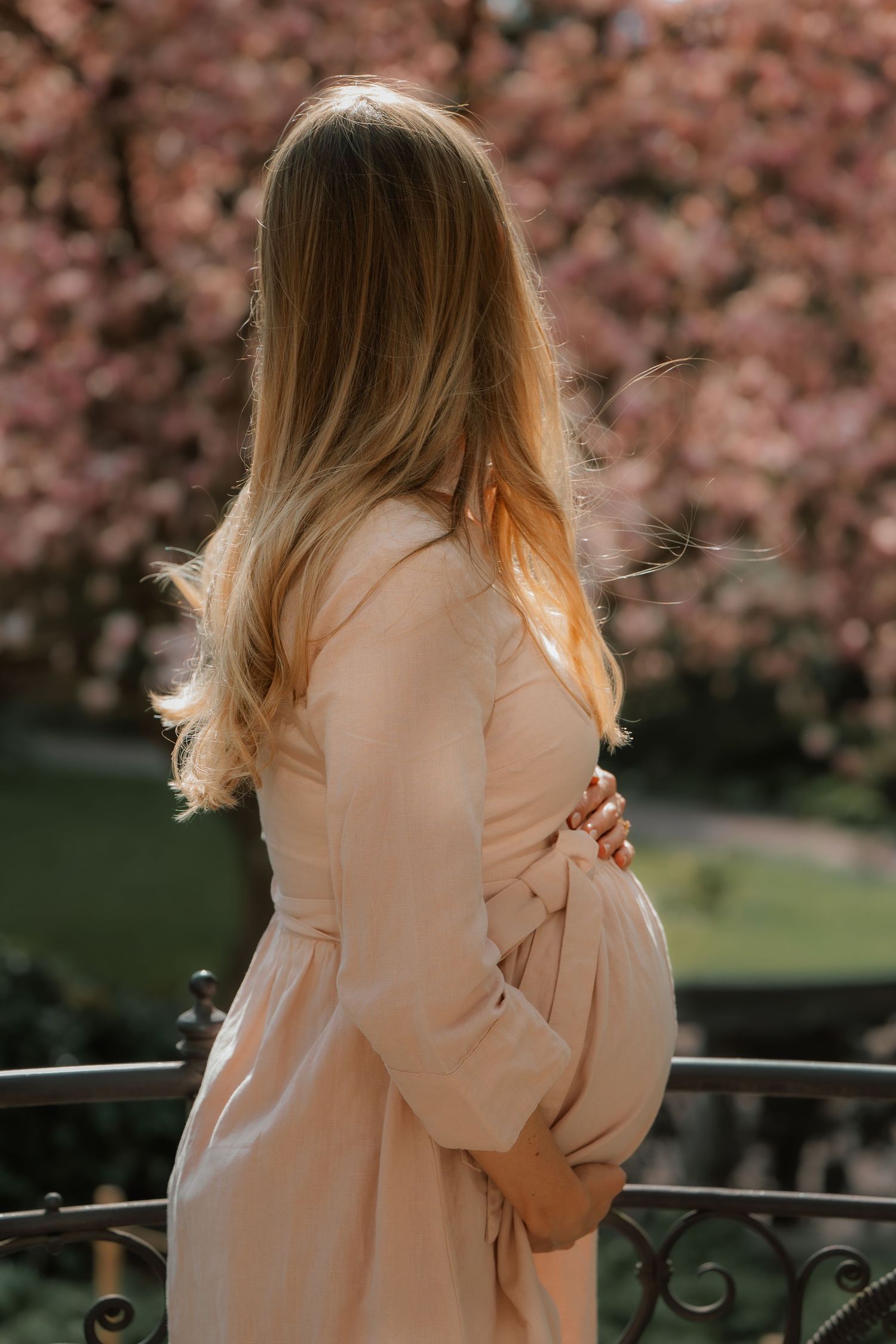 Pregnant woman in beige dress cradling baby bump outdoors near blooming pink cherry blossom trees in spring.