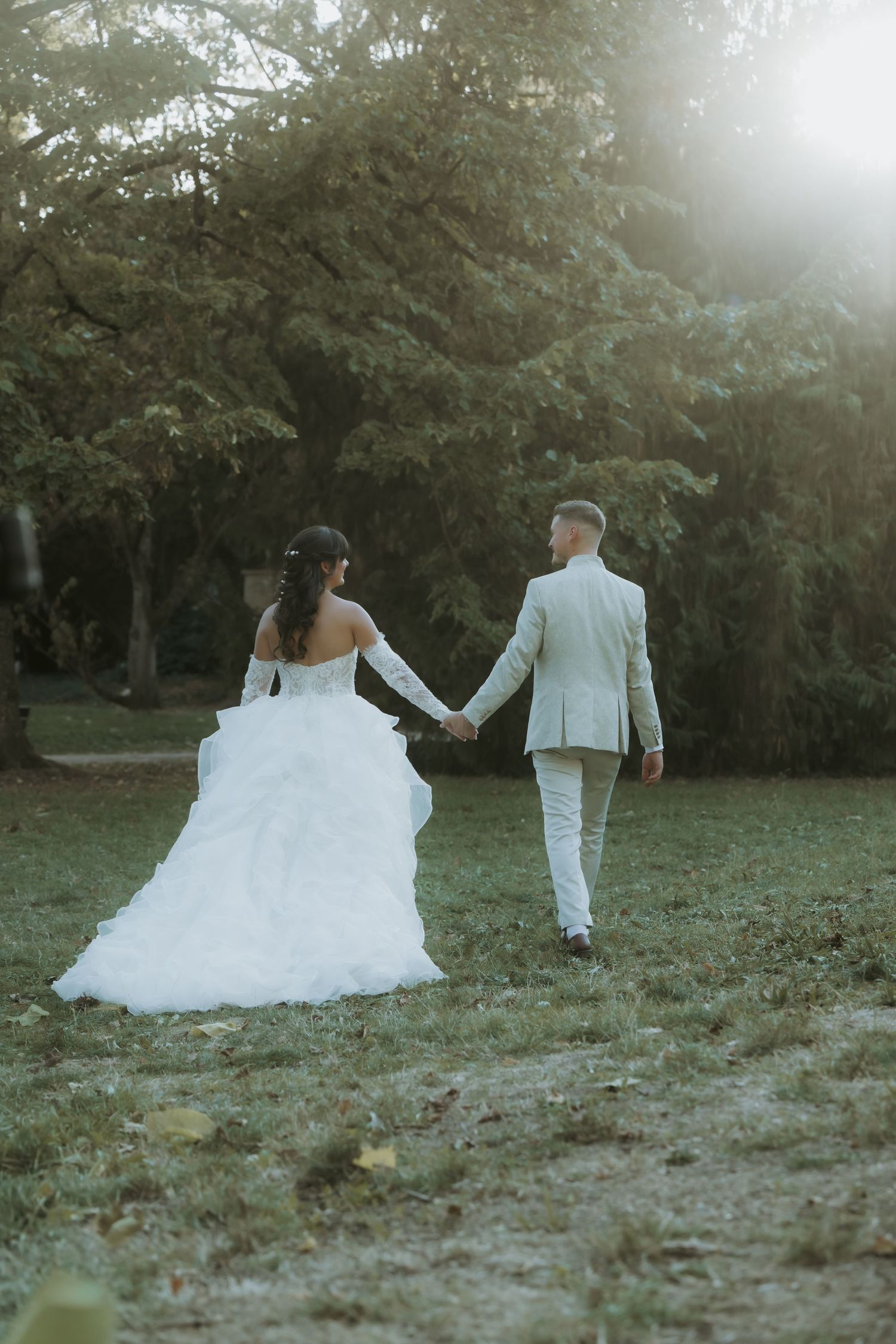 A bride in a white gown and groom in a light suit walk hand in hand through a lush green park at sunset.