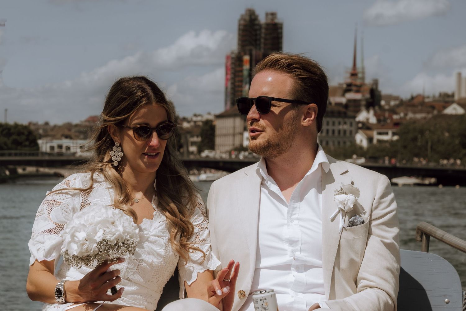 Bride and groom in sunglasses sitting together on a boat with a city skyline and river in the background.