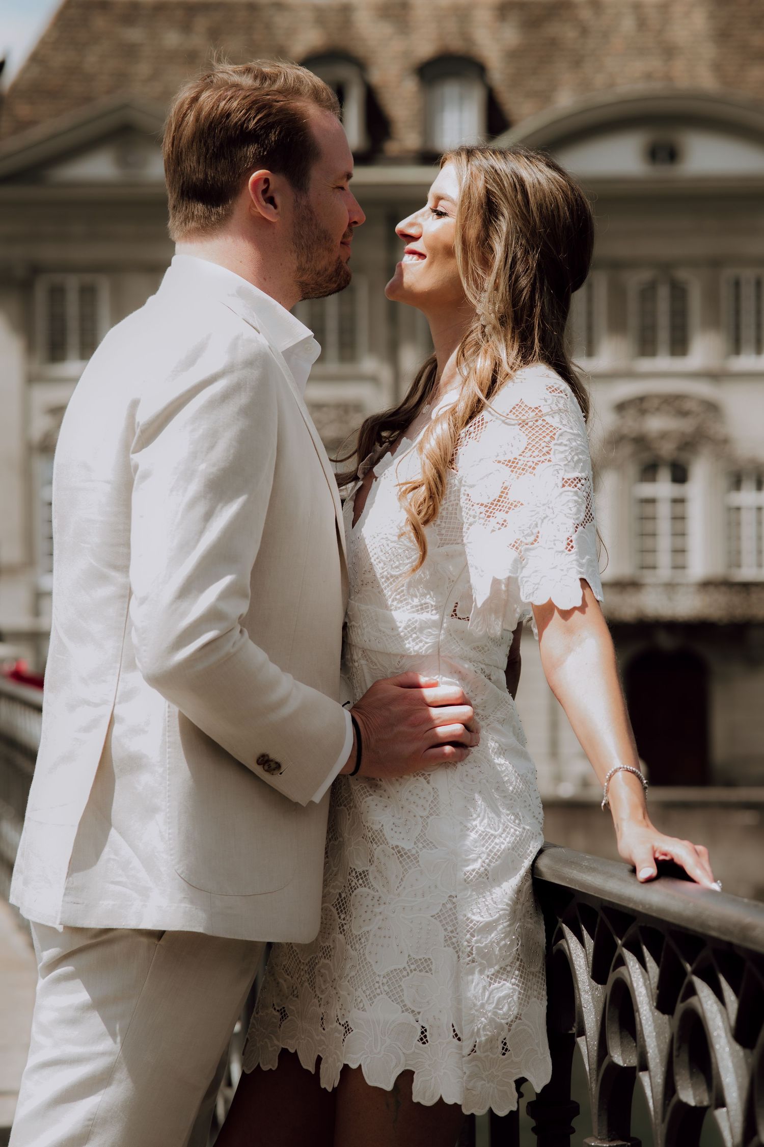 A couple in white wedding attire share a romantic moment on a balcony with a historic building backdrop.