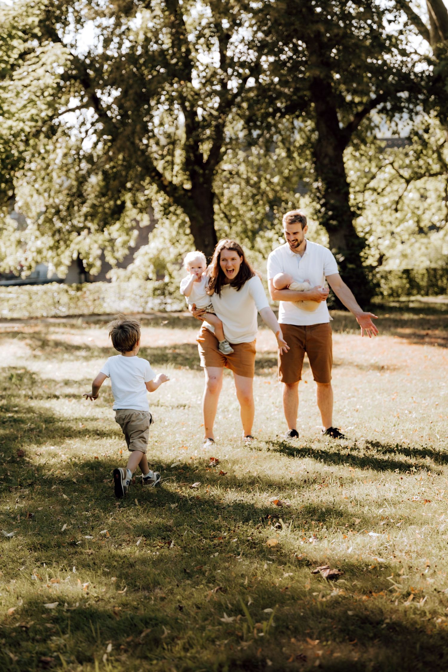 Family playing outdoors in sunny park, parents welcoming toddler running toward them while holding baby.