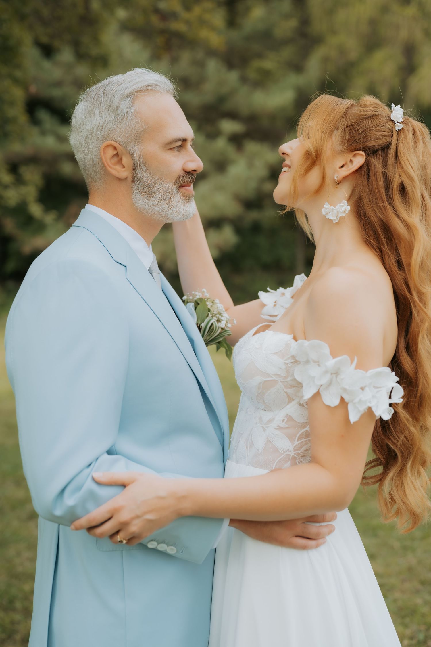 A bride in a white floral gown and groom in a light blue suit share a romantic embrace outdoors.