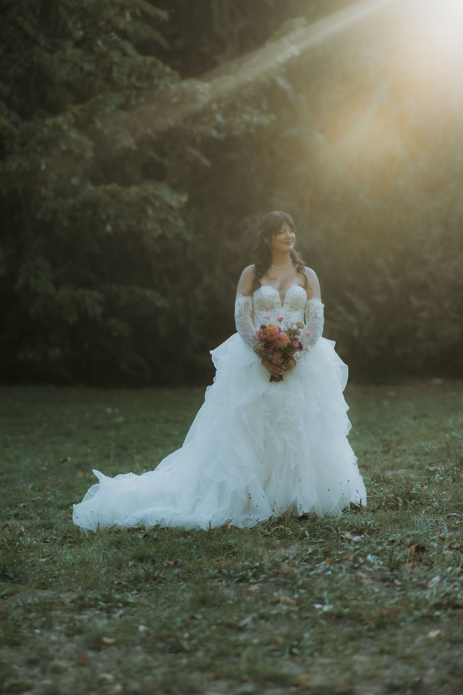 Bride in white ruffled gown holding pink bouquet, standing in sunlit garden with golden hour light.