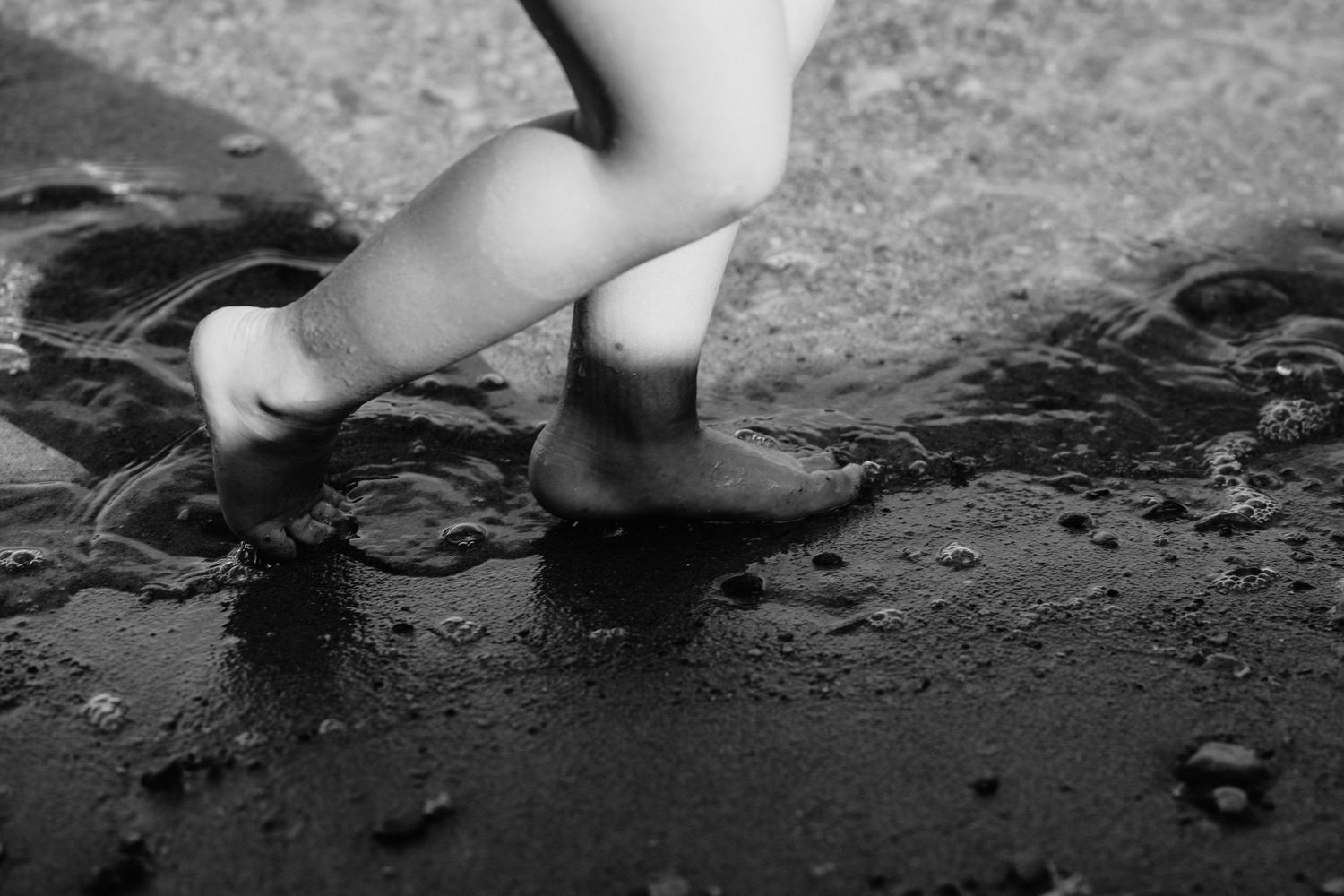 Black and white photo of a child's bare feet splashing in a rain puddle on wet pavement.