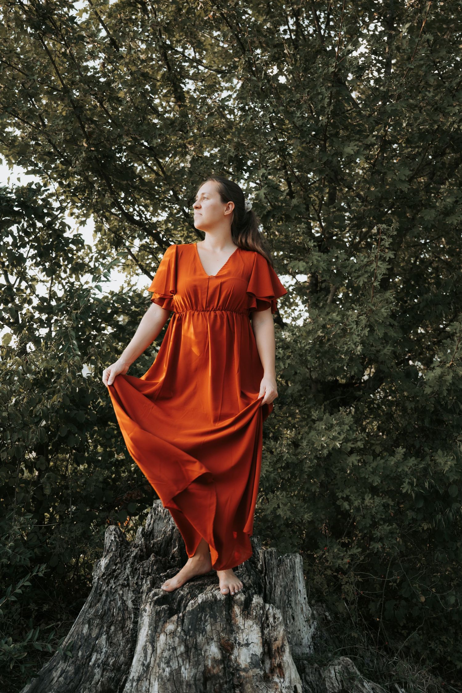 Woman in flowing orange dress standing barefoot on tree stump surrounded by lush green trees.