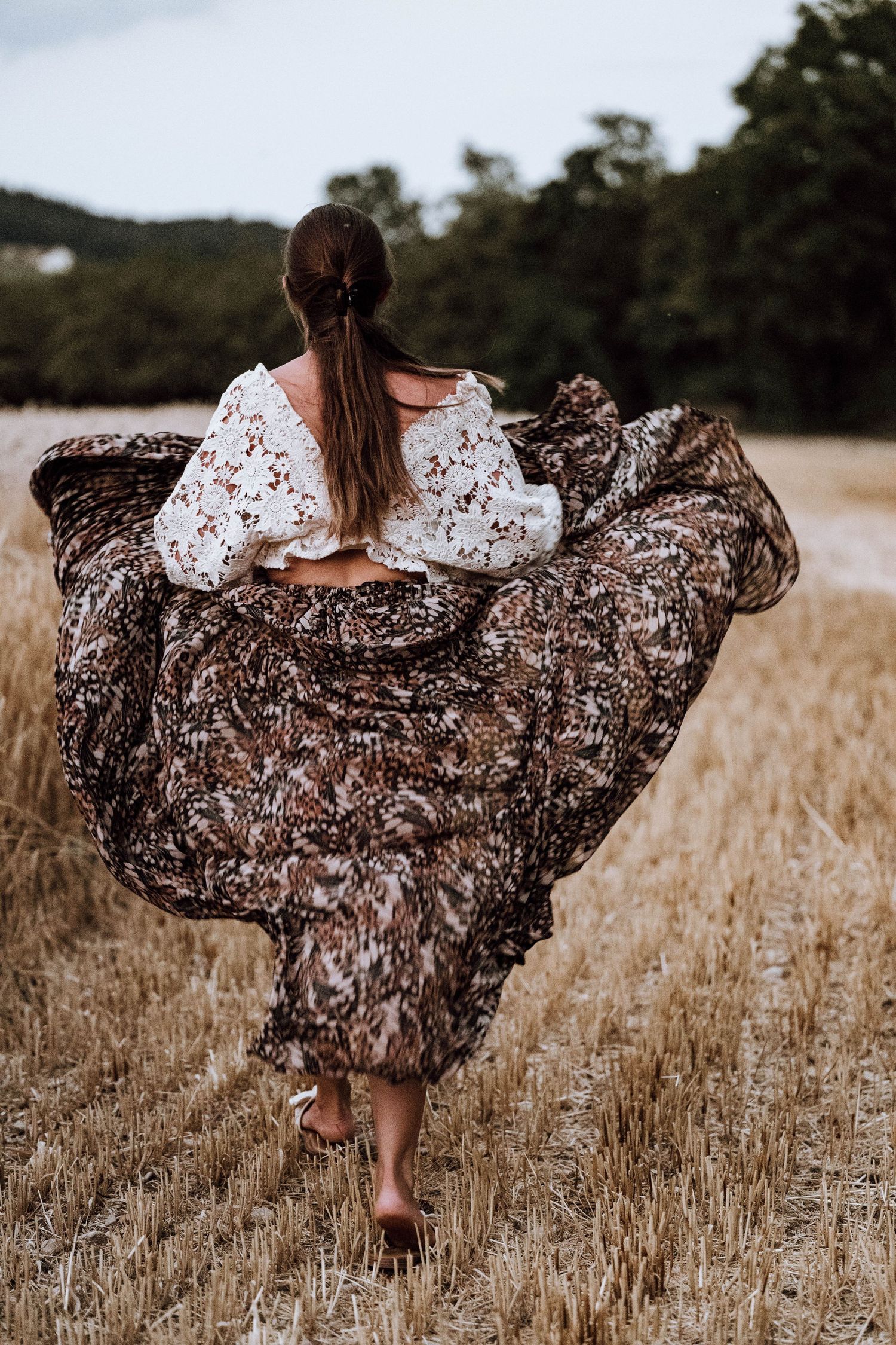 Woman in floral skirt and lace top walking through golden wheat field, skirt billowing in the wind.