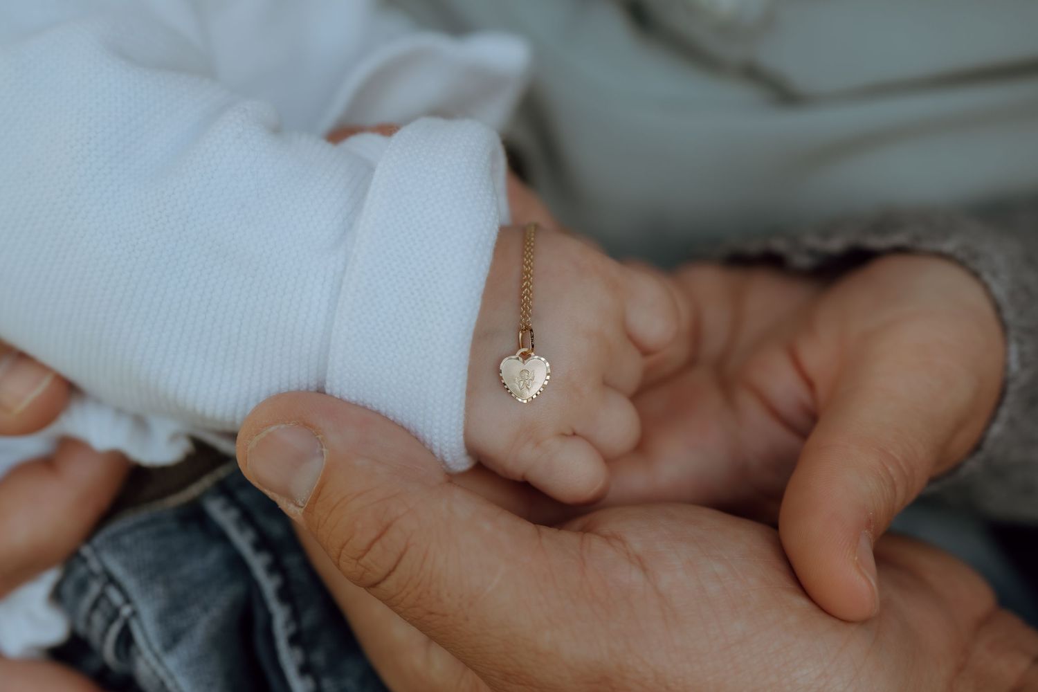 A newborn baby wearing a delicate gold heart bracelet rests their tiny hand in an adult's gentle hands.