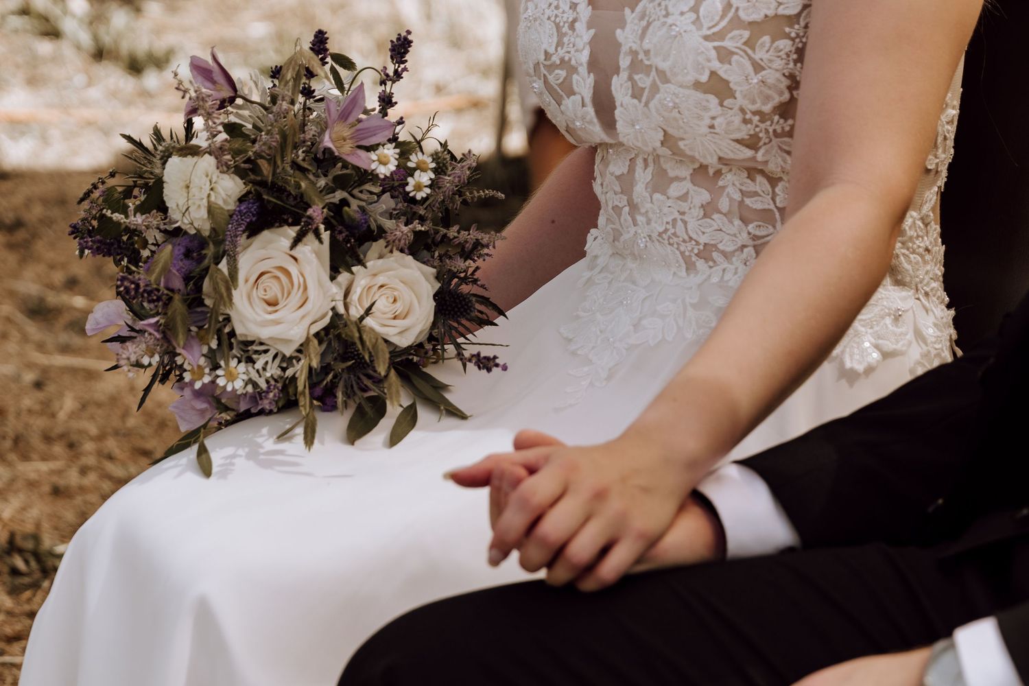 Bride and groom sitting together, holding hands, with a beautiful purple and white wedding bouquet.