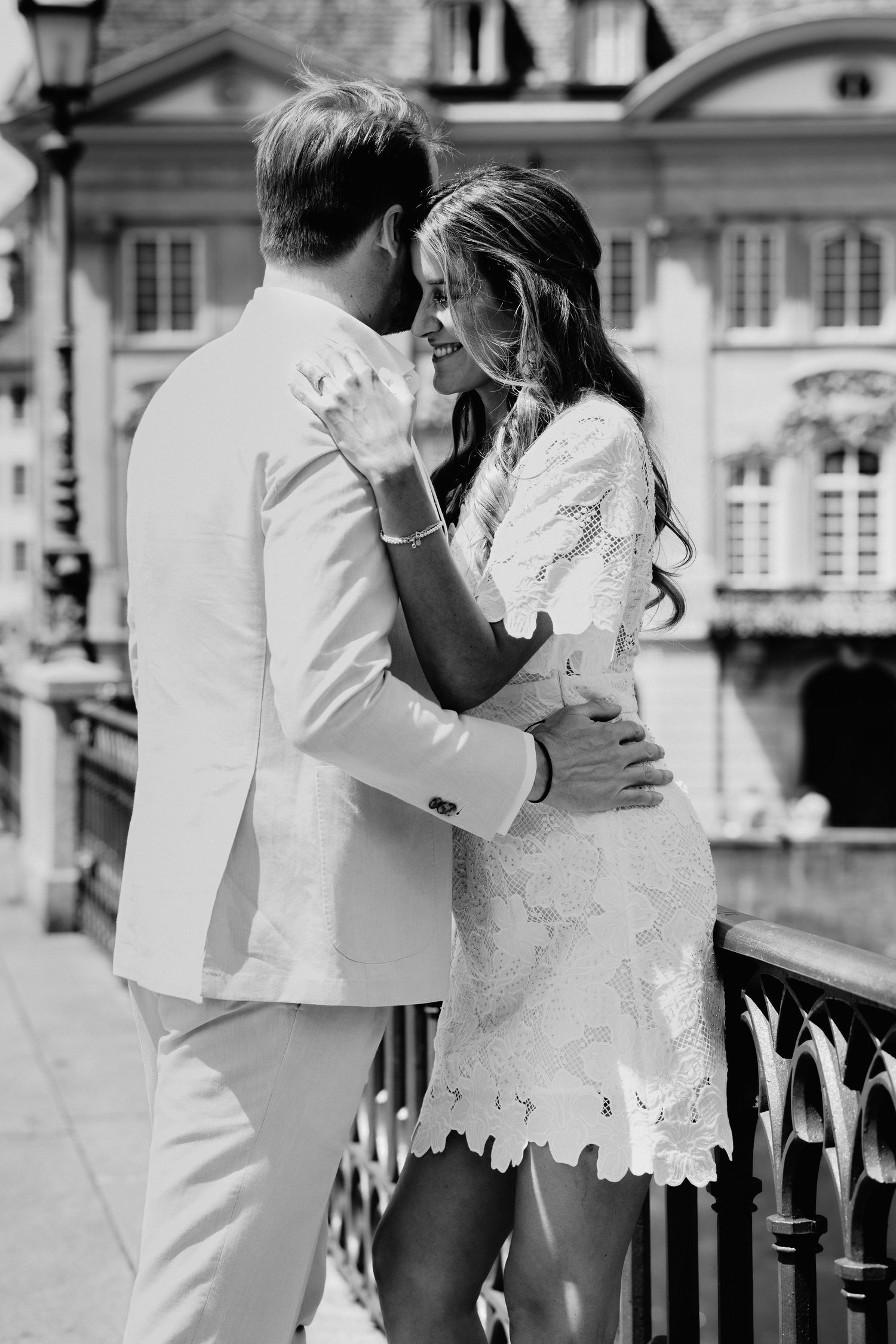 Black and white photo of a couple embracing on a bridge, woman in lace dress smiling at man in white suit.