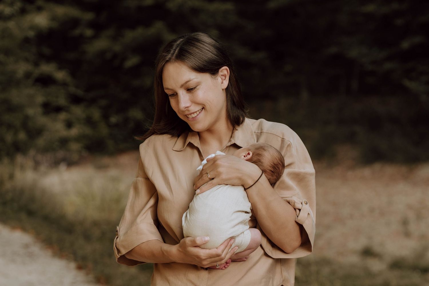 A smiling mother tenderly holds her newborn baby outdoors, wearing a beige shirt with a forest backdrop.