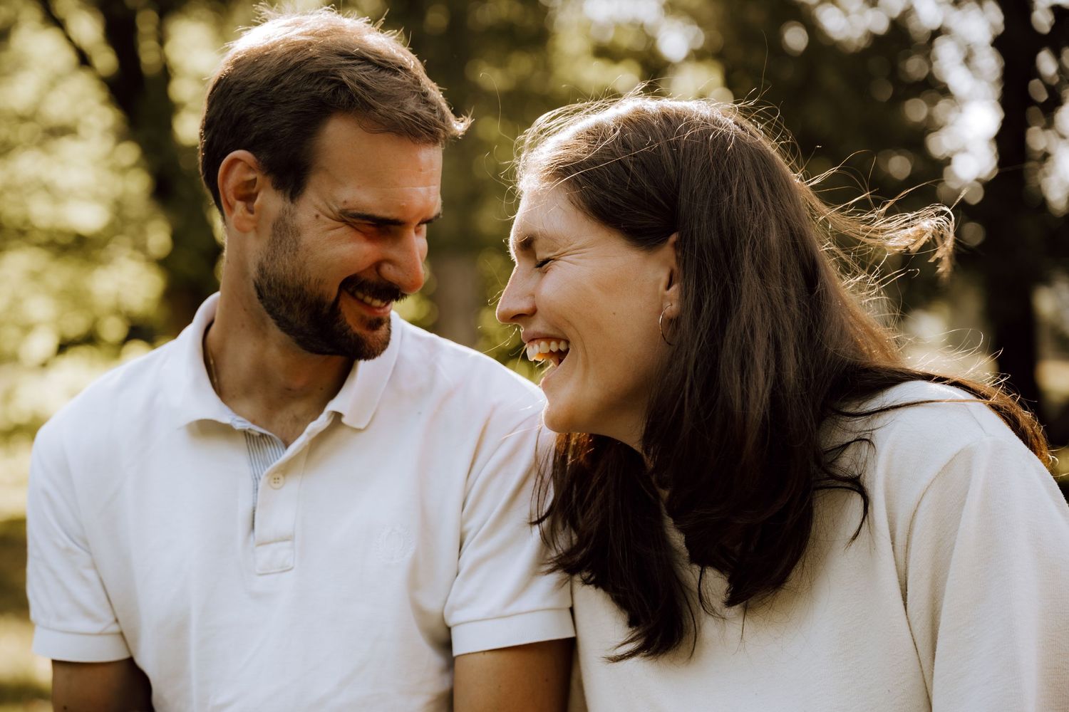 A smiling couple in matching white polo shirts share a joyful moment outdoors in a sunlit park.