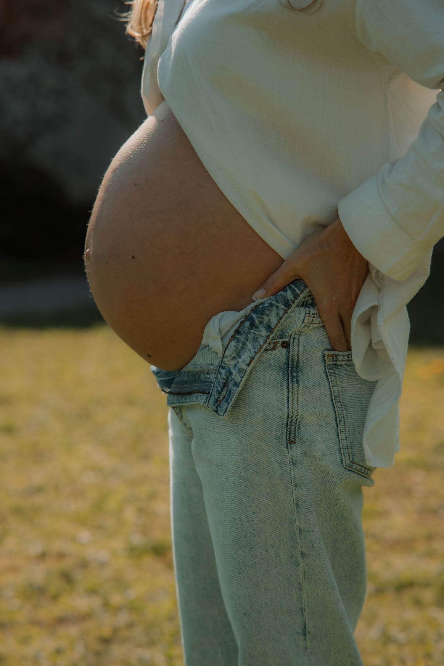 Pregnant woman in white shirt and light blue jeans outdoors, showcasing baby bump in natural sunlight.