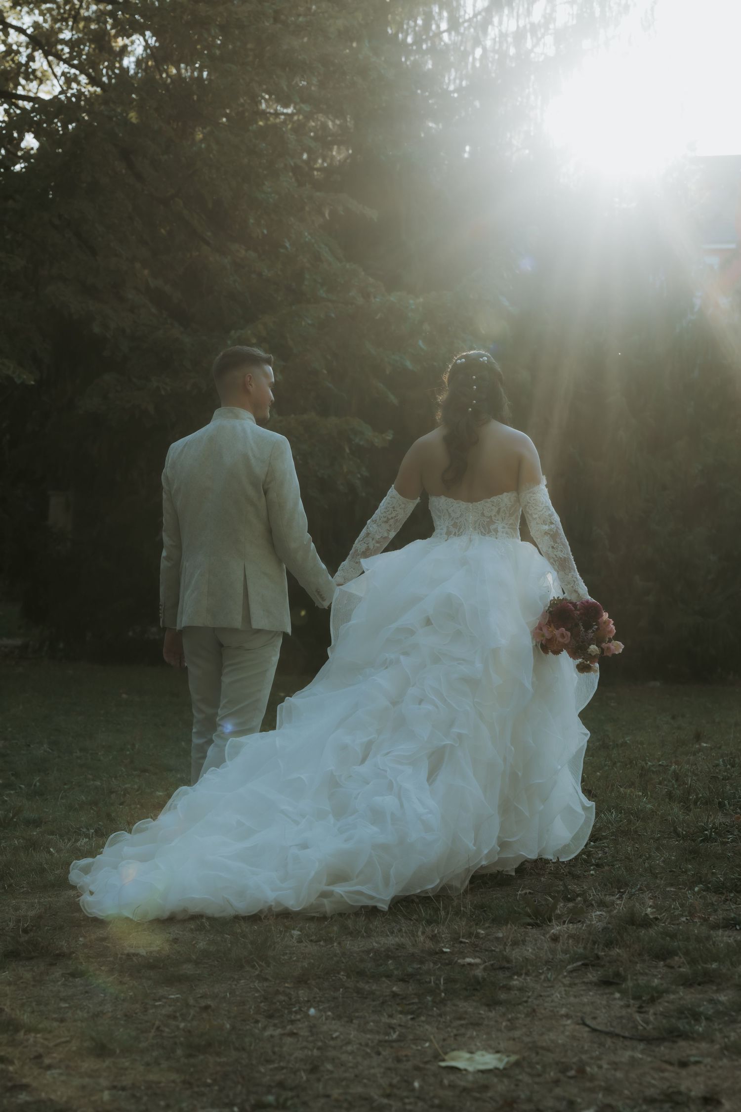 Bride in white gown and groom in tan suit walk hand-in-hand toward sunlit trees in a romantic outdoor wedding photo.