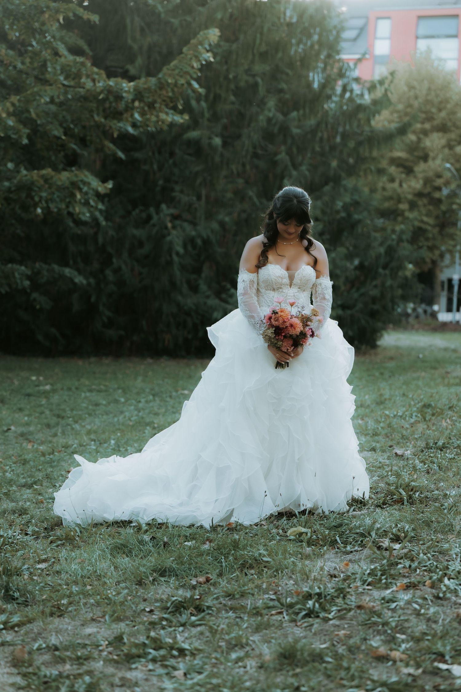 Bride in a white ruffled ball gown holding a colorful bouquet, standing on green grass with trees in the background.