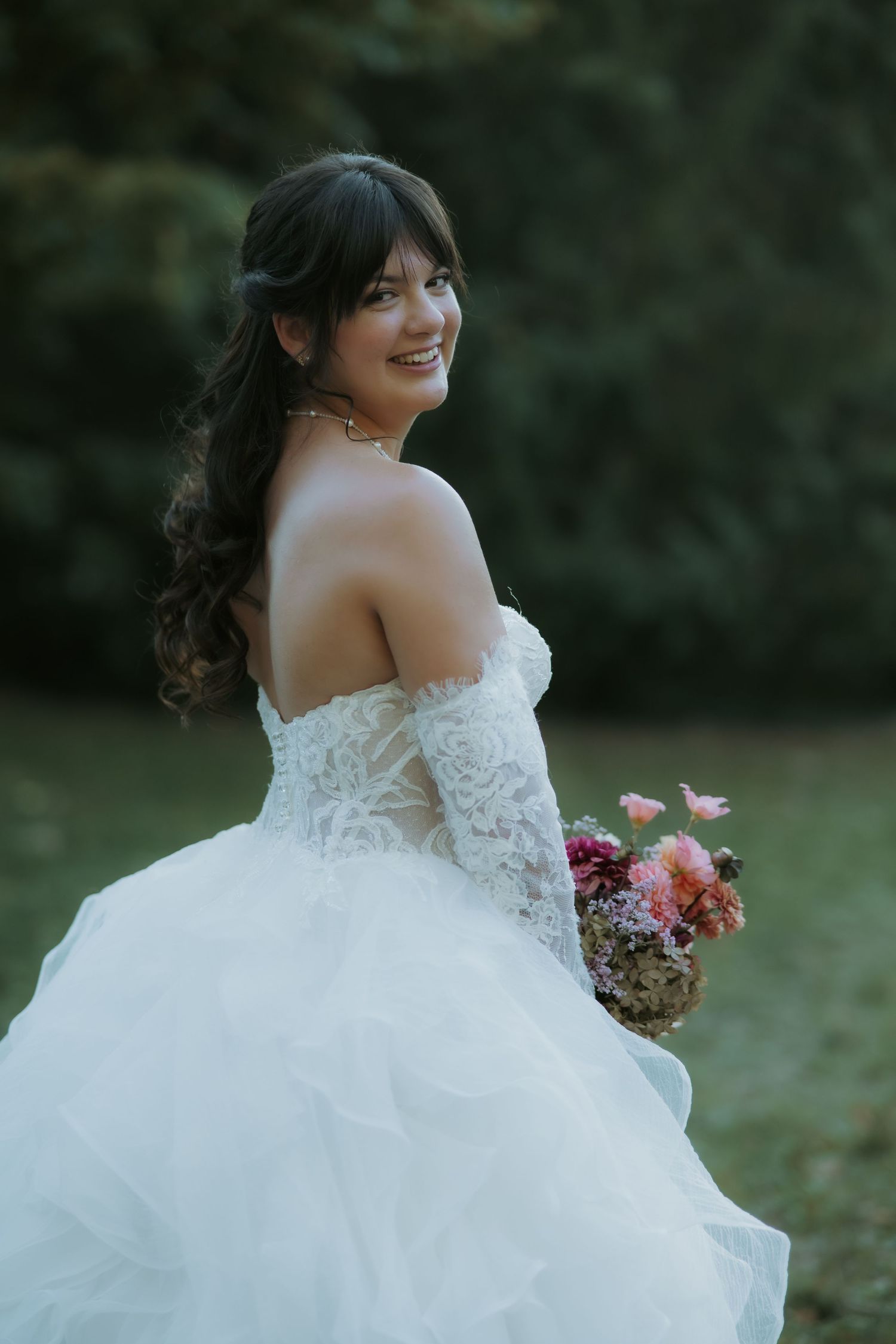 Smiling bride in off-shoulder lace wedding gown holding pink bouquet, posing outdoors with green trees background.