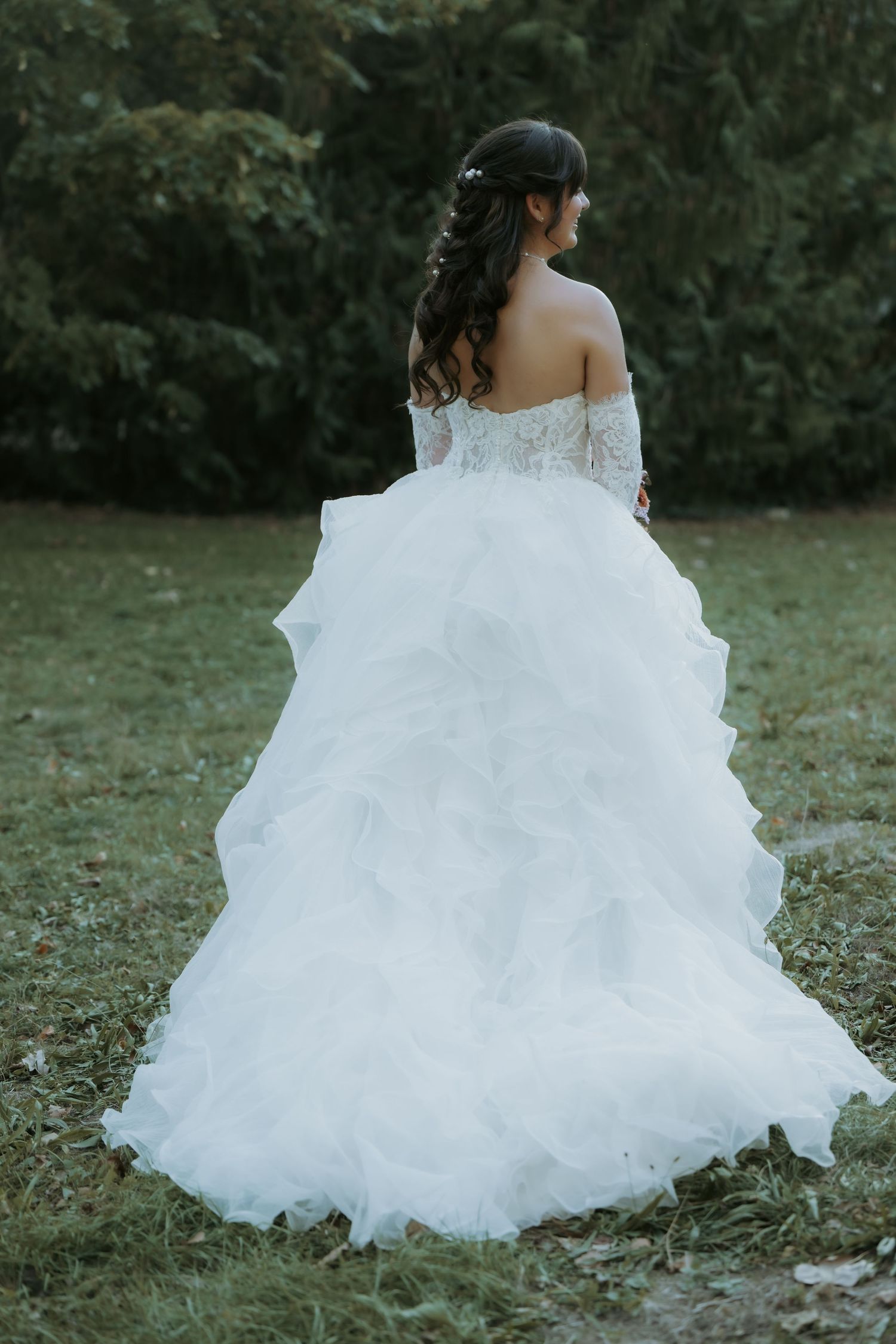 Bride in white off-shoulder ball gown with ruffled skirt and lace bodice, standing in garden, viewed from behind.