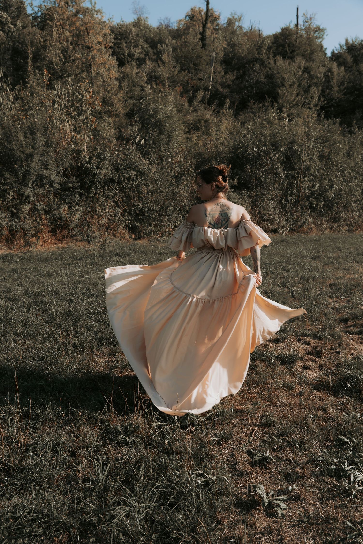 Woman in flowing peach gown twirling in a sunlit meadow with lush green trees in the background.