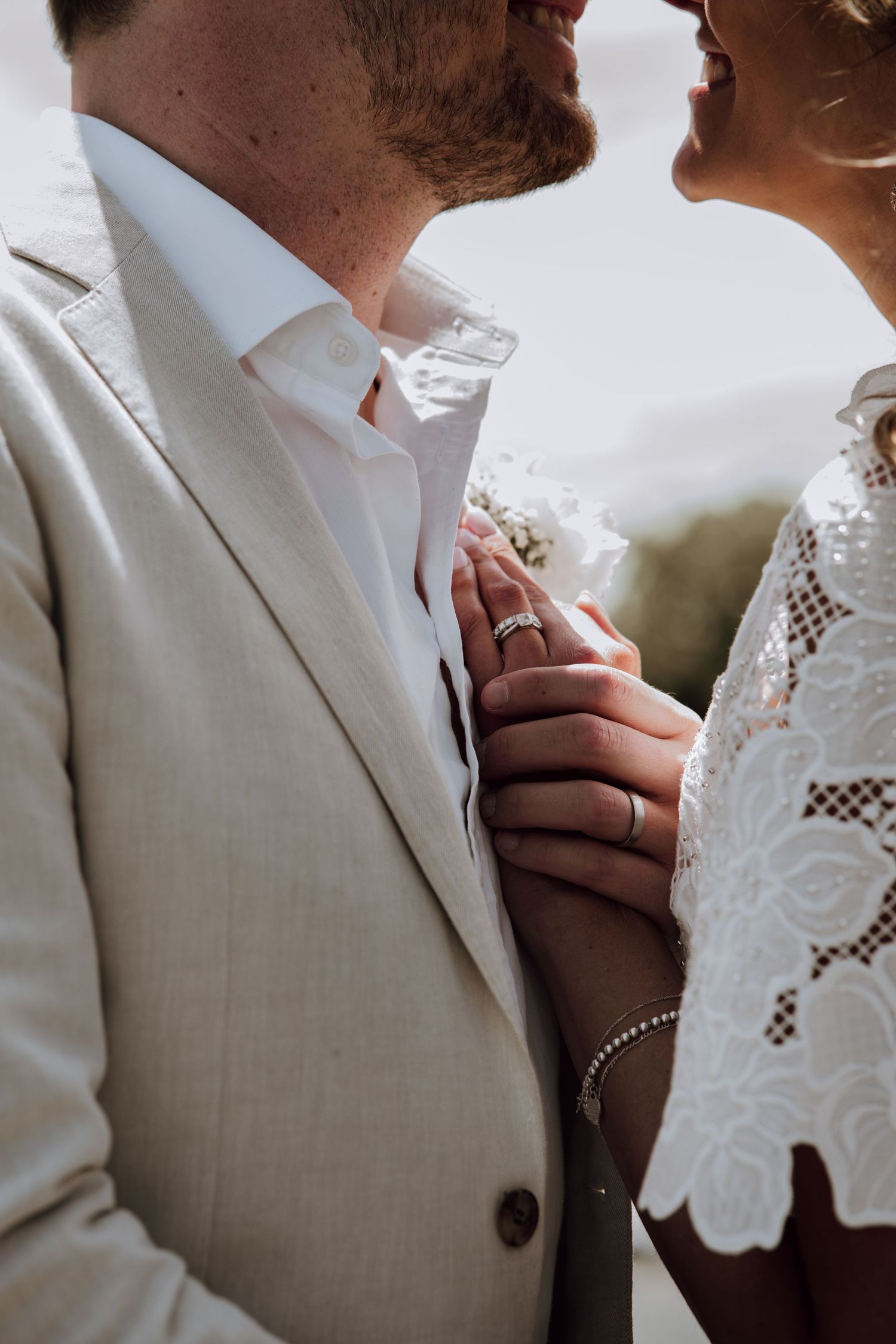 Close-up of wedding couple in lace dress and beige suit, showing rings and boutonniere during ceremony.