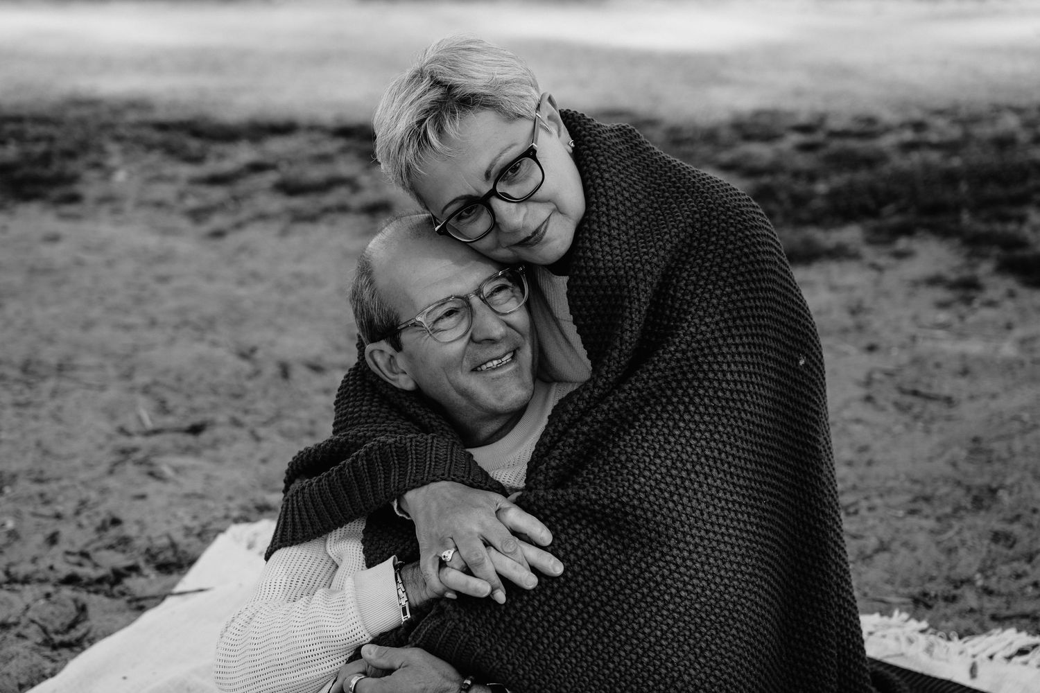 Elderly couple sharing a warm embrace outdoors, wrapped in a blanket, smiling joyfully in a black and white photo.