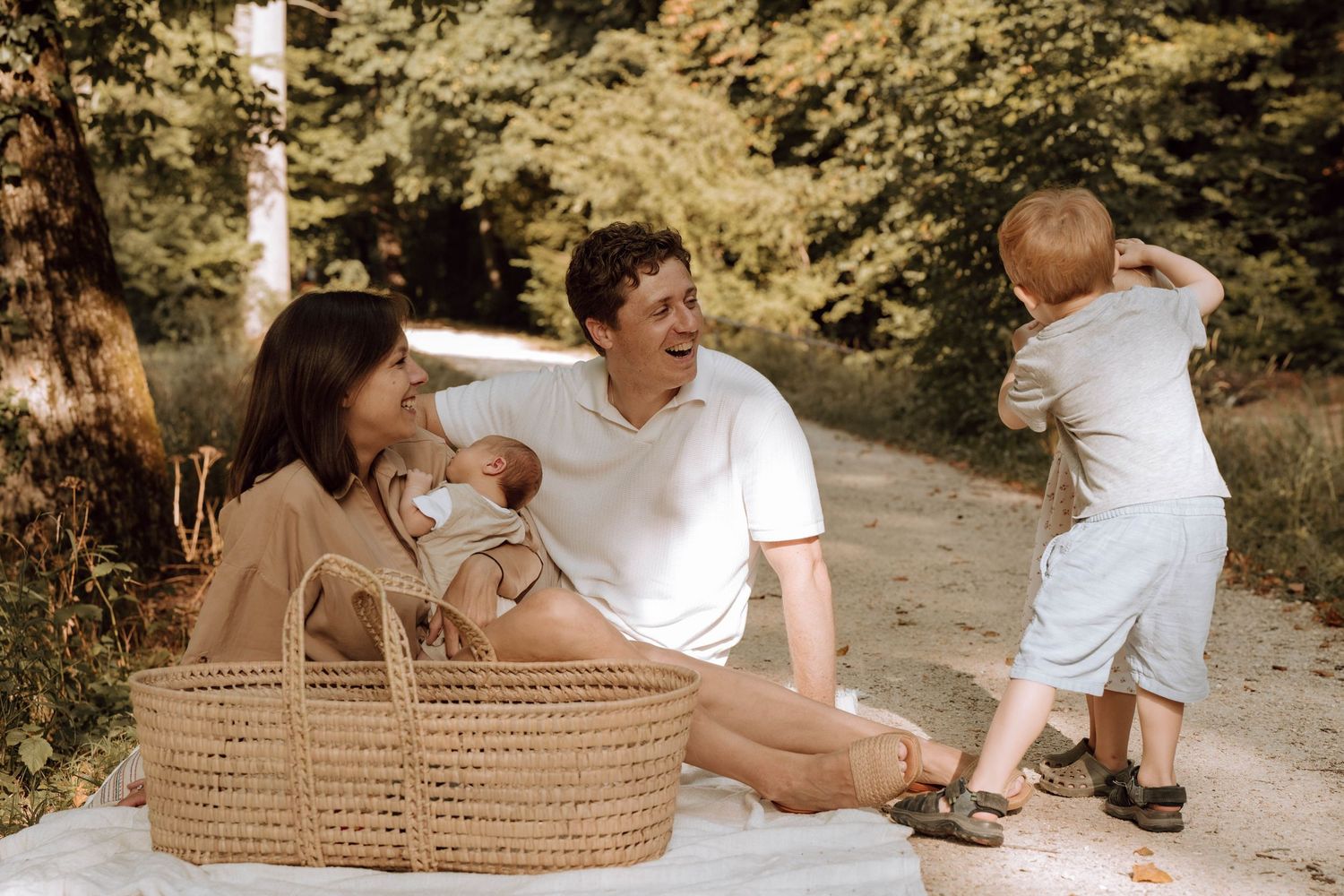 Family enjoying a sunny outdoor picnic on a blanket with a wicker basket, baby, and toddler in a lush green park.