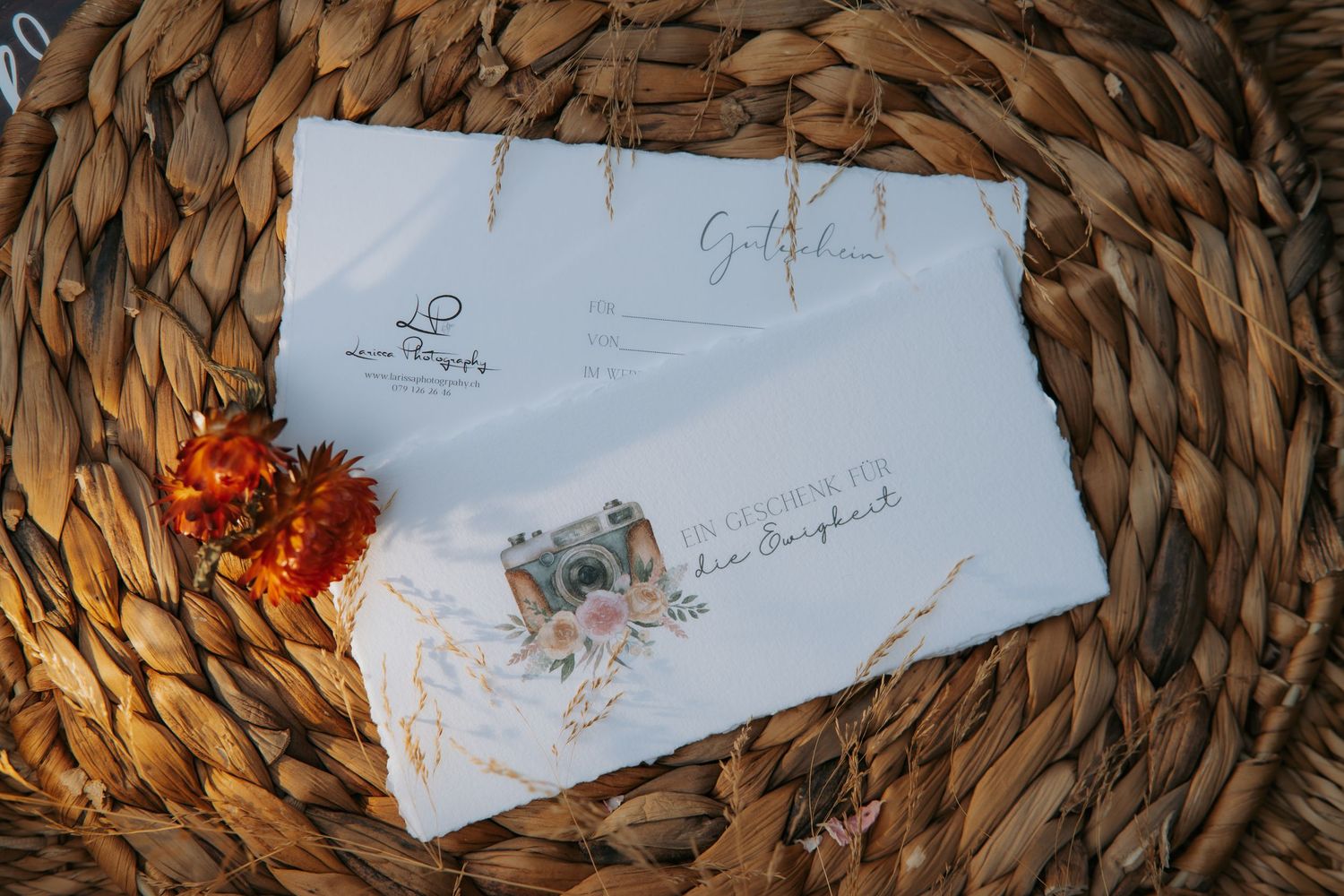 White envelopes with elegant calligraphy and a vintage camera stamp rest on a woven rattan surface with dried orange flowers.